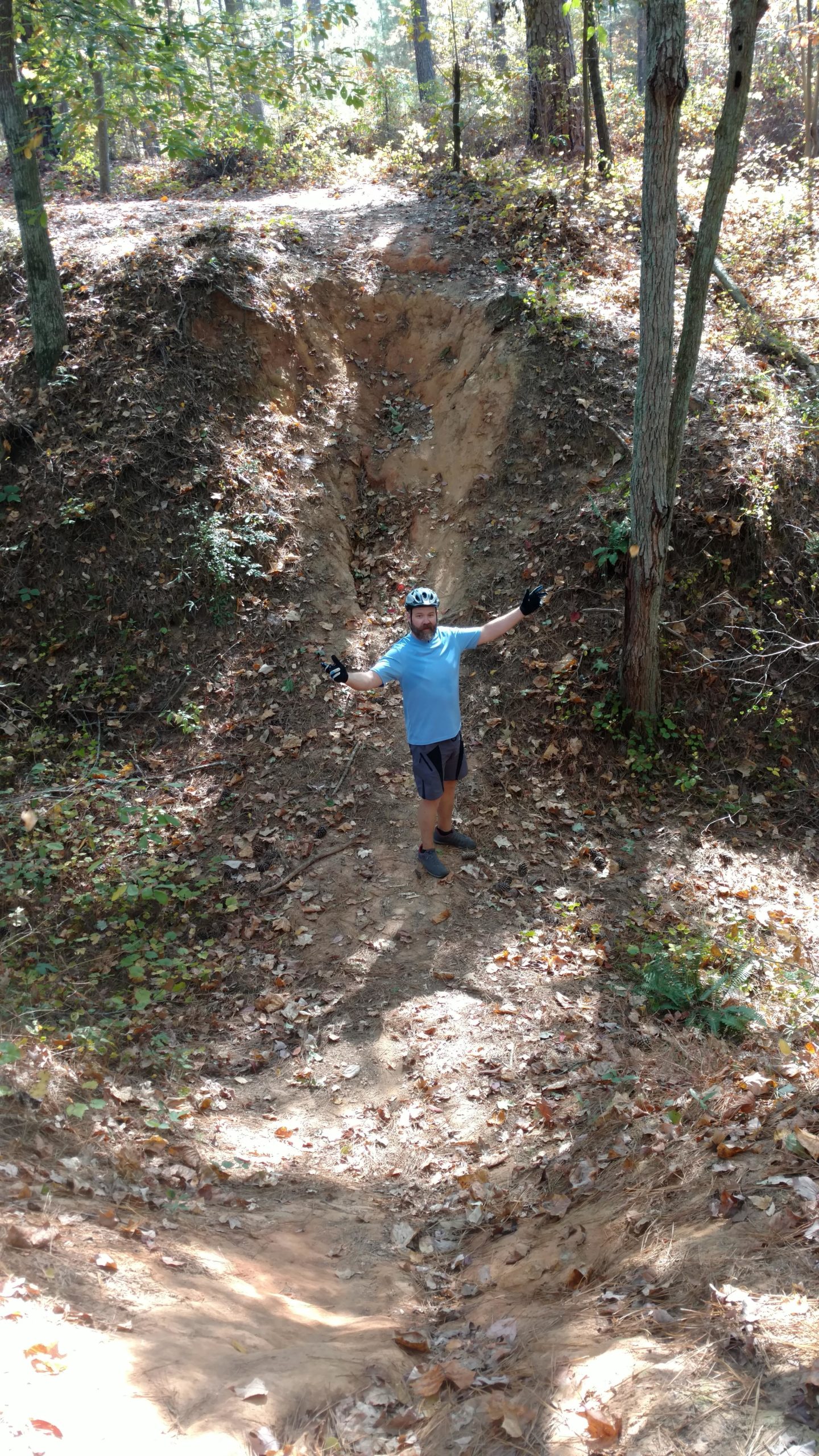 A person in a blue shirt and helmet stands in a dirt path surrounded by trees, with arms raised, celebrating or posing in an outdoor setting. The ground is covered with fallen leaves, and the scene is illuminated by sunlight filtering through the trees. Fort Yargo State Park mountain bike trail.