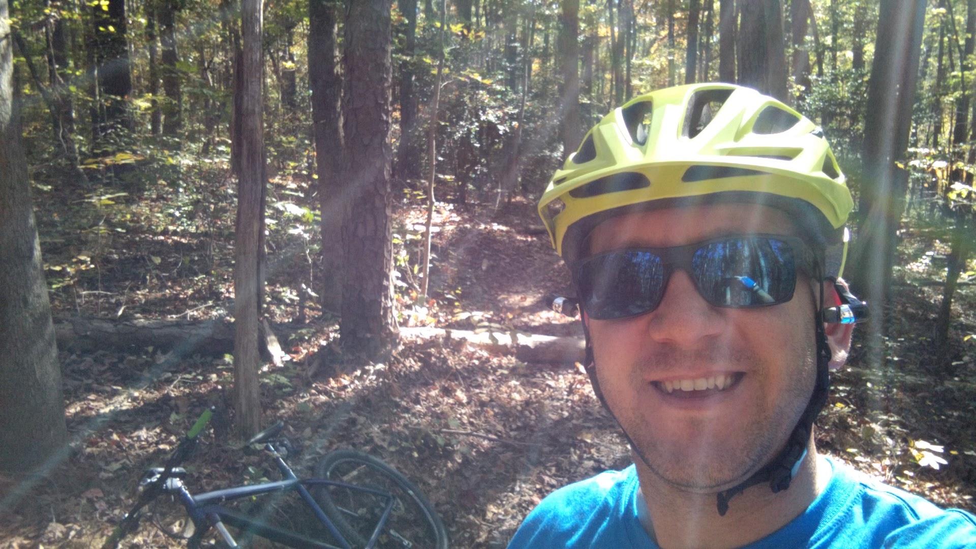 A smiling individual in a bright yellow cycling helmet and sunglasses stands in a wooded area, with a mountain bike resting nearby. Sunlight filters through the trees, casting a warm glow on the scene. The background features leafy ground cover and tree trunks, creating a serene outdoor atmosphere. San-lee Park mountain bike trail.