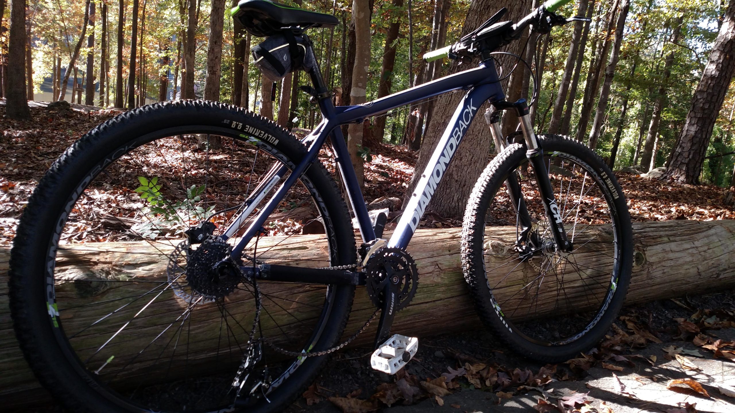 A mountain bike leaning against a log in a wooded area, surrounded by fall foliage. The bike is dark blue with white branding and features thick tires suitable for off-road cycling. Sunlight filters through the trees, illuminating the scene. San-lee Park mountain bike trail.