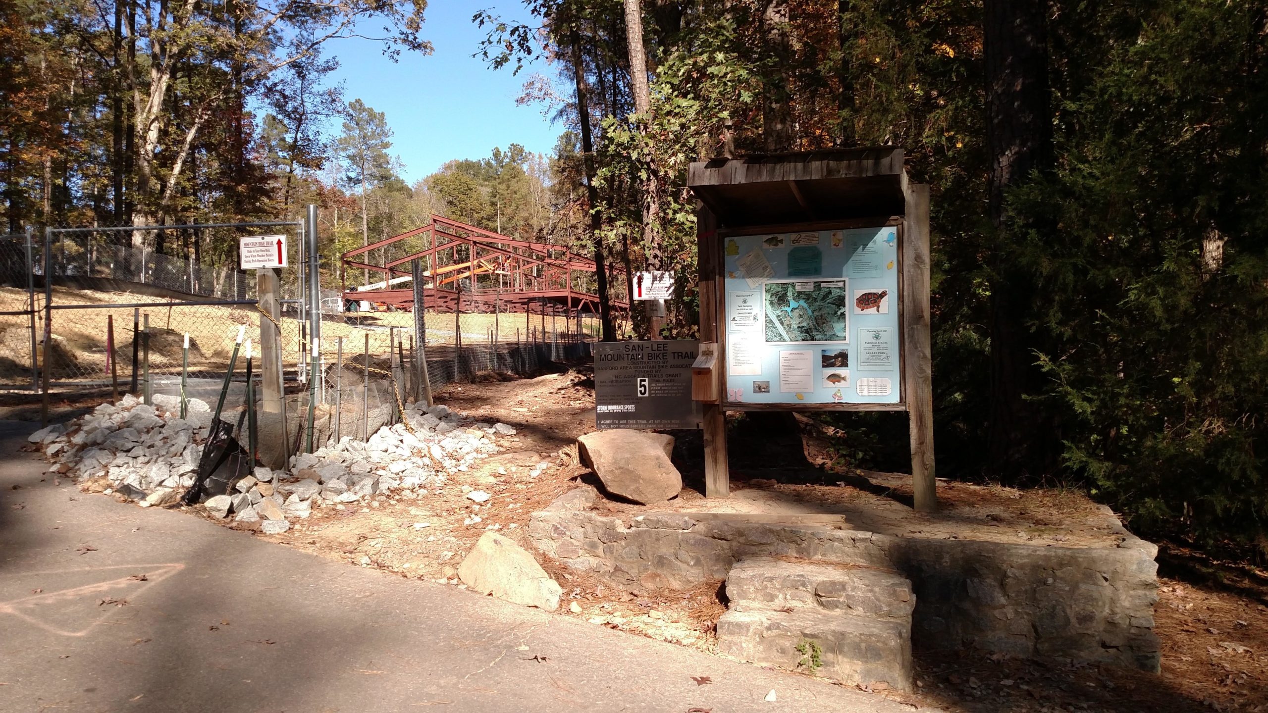 Sign at the entrance of the San-Lee Mountain Bike Trail, featuring an informational board with maps and details about the trail, surrounded by trees in a natural setting. A construction site is visible in the background, indicating ongoing improvements. The path is lined with rocks and a fence. San-lee Park mountain bike trail.