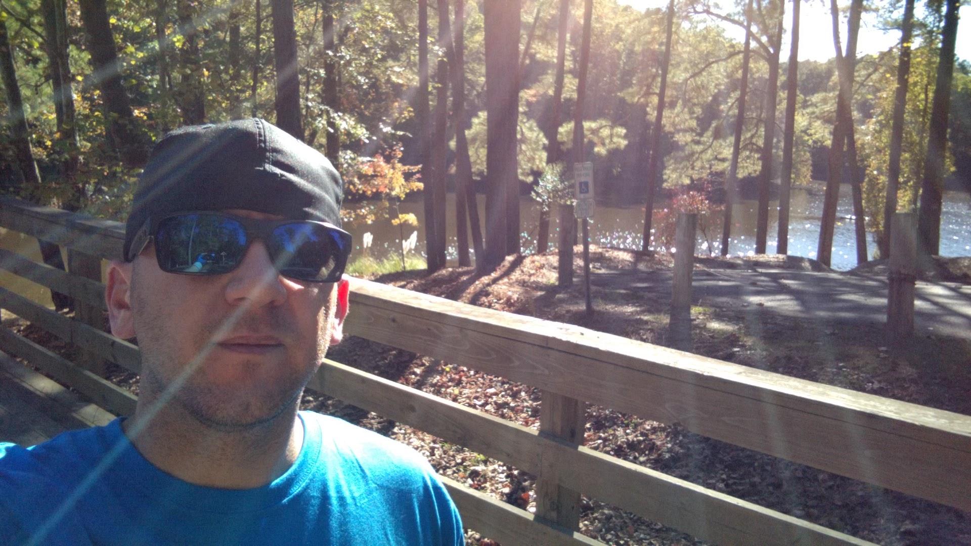 A man wearing sunglasses and a black cap stands in front of a wooden railing, with a scenic view of a lake and trees in the background. Sunlight filters through the foliage, creating a bright and serene atmosphere. San-lee Park mountain bike trail.