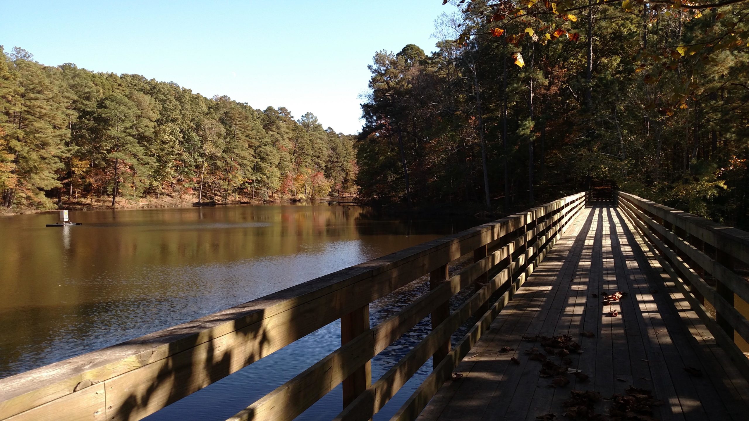 A scenic view of a wooden bridge extending across a calm lake, surrounded by lush green trees showing hints of autumn colors. The sunlight casts long shadows on the bridge's planks, and a small sailboat can be seen gliding on the water in the distance. San-lee Park mountain bike trail.