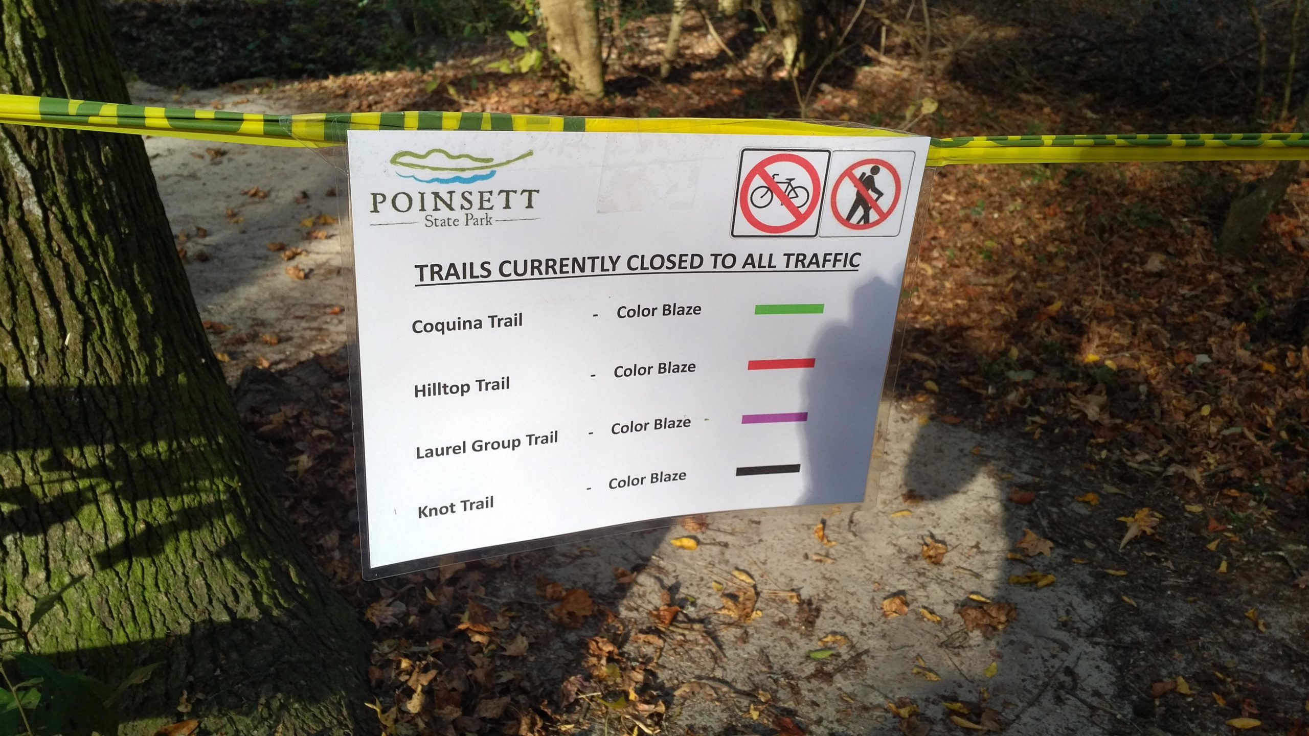 A sign at Poinsett State Park indicating that all trails are currently closed to traffic. The sign lists the trails—Coquina Trail, Hilltop Trail, Laurel Group Trail, and Knot Trail—along with their corresponding color blazes. The sign is surrounded by yellow caution tape and is situated in a wooded area with fallen leaves on the ground. The Knot mountain bike trail.