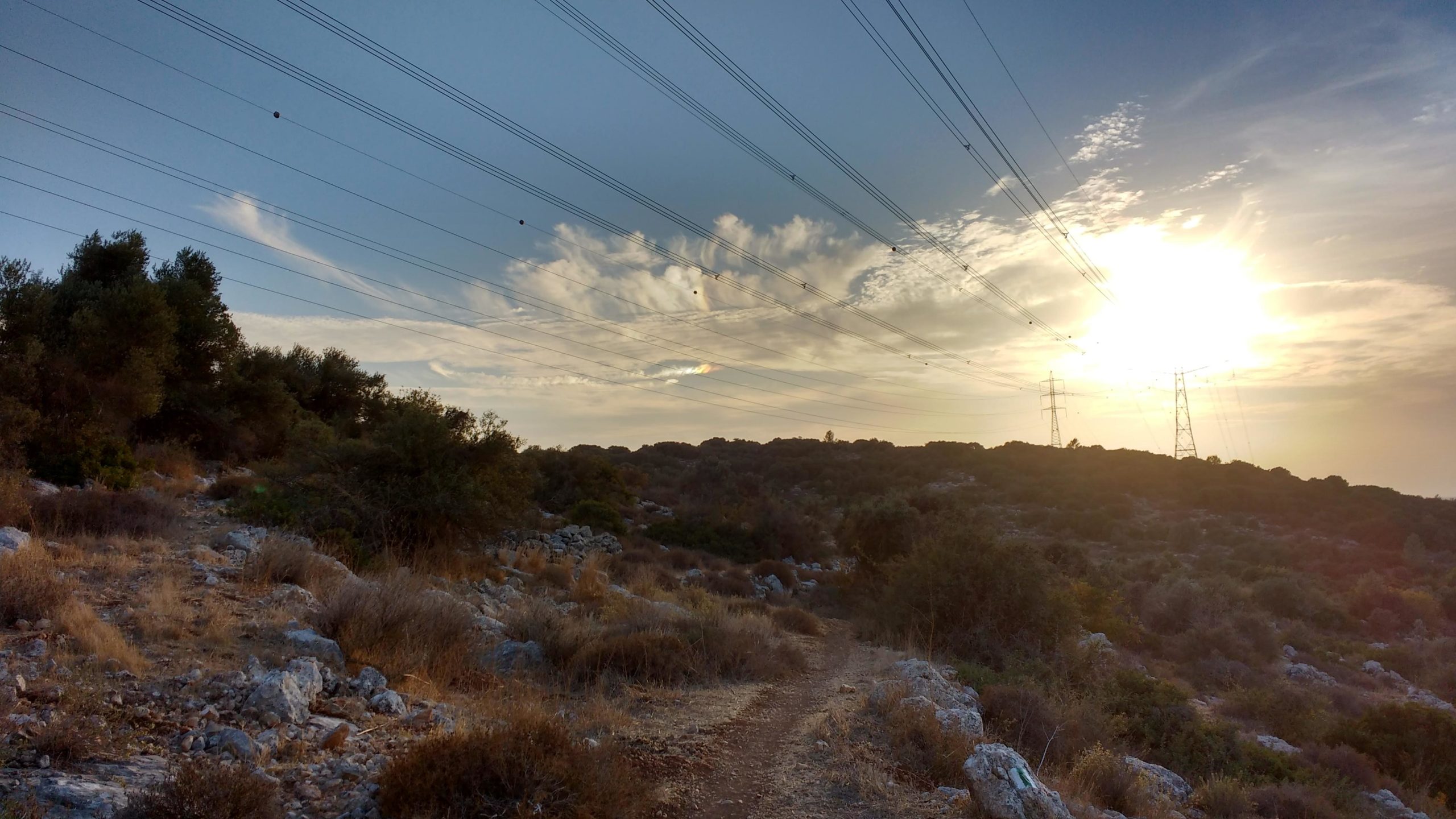 A scenic landscape featuring a dirt path winding through rocky terrain, flanked by vegetation and shrubs. In the background, tall power lines stretch across the sky, silhouetted against a sunset filled with soft clouds and warm light. Zechariah singletrack mountain bike trail.