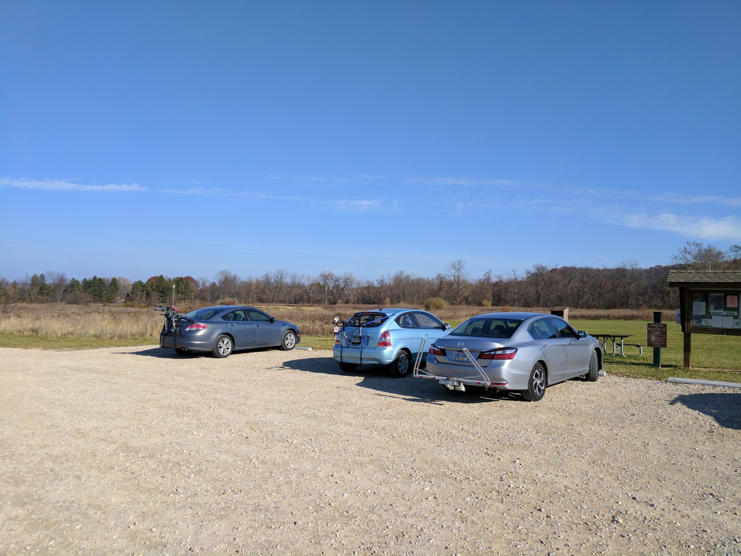Three parked cars are visible in a gravel parking area surrounded by open fields and trees under a clear blue sky. In the background, there is a signboard and a picnic table. The scene suggests a recreational area, likely popular for outdoor activities. Chestnut Ridge mountain bike trail.