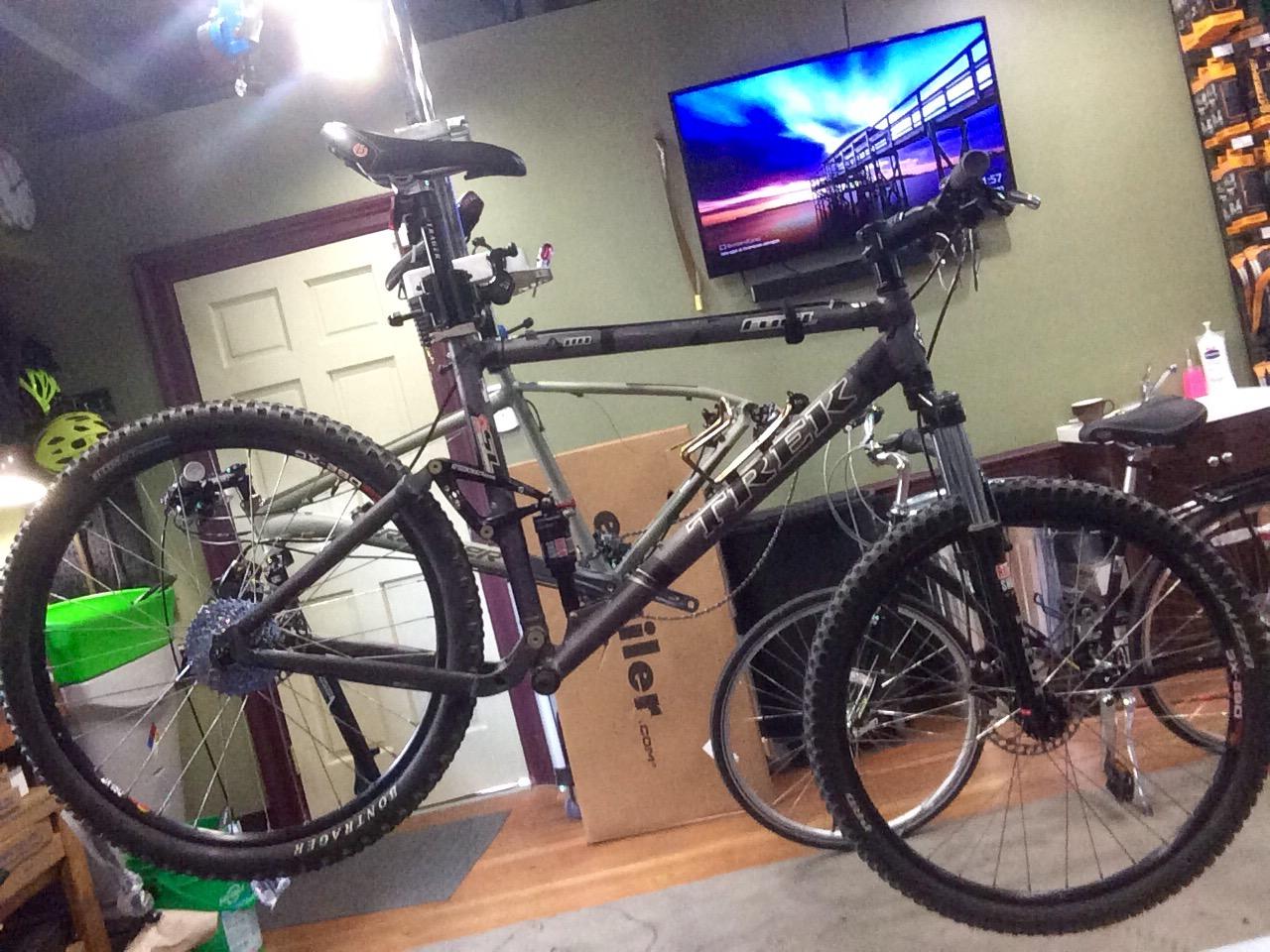Trek Fuel EX: A black Trek mountain bike is suspended in a garage, with its rear wheel visible. The background includes a door, a television displaying a scenic image, and various items related to cycling, including another bike and storage boxes. The workspace is well-lit, highlighting the bike's features and surrounding environment.