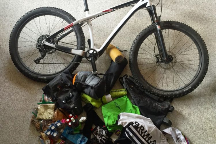 A top-down view of a mountain bike alongside a variety of cycling gear and supplies spread out on a carpeted surface. The bike features thick tires and a sleek frame, while the assortment includes clothing, a helmet, tools, snacks, and various cycling accessories, indicating preparation for an outdoor biking adventure.