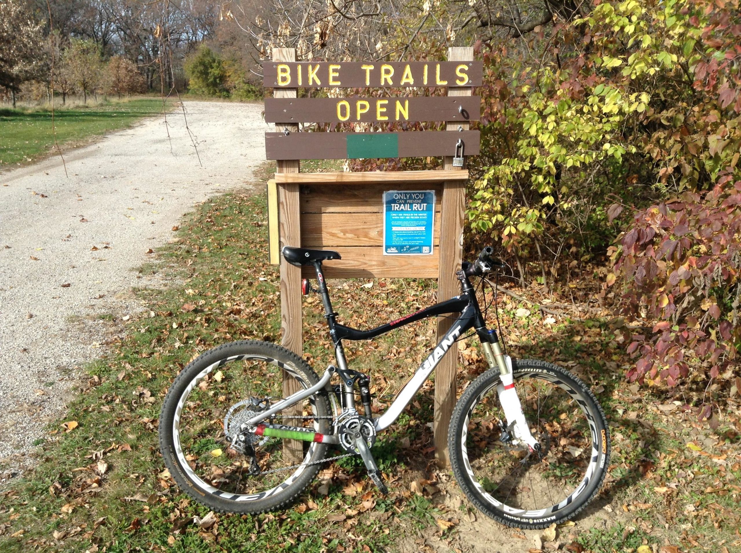A mountain bike leaning against a sign that reads "Bike Trails Open." The sign is wooden and displays a blue poster with additional information. Surrounding the sign are colorful autumn leaves, and a gravel path is visible leading into a wooded area. Illiniwek Forest Preserve mountain bike trail.