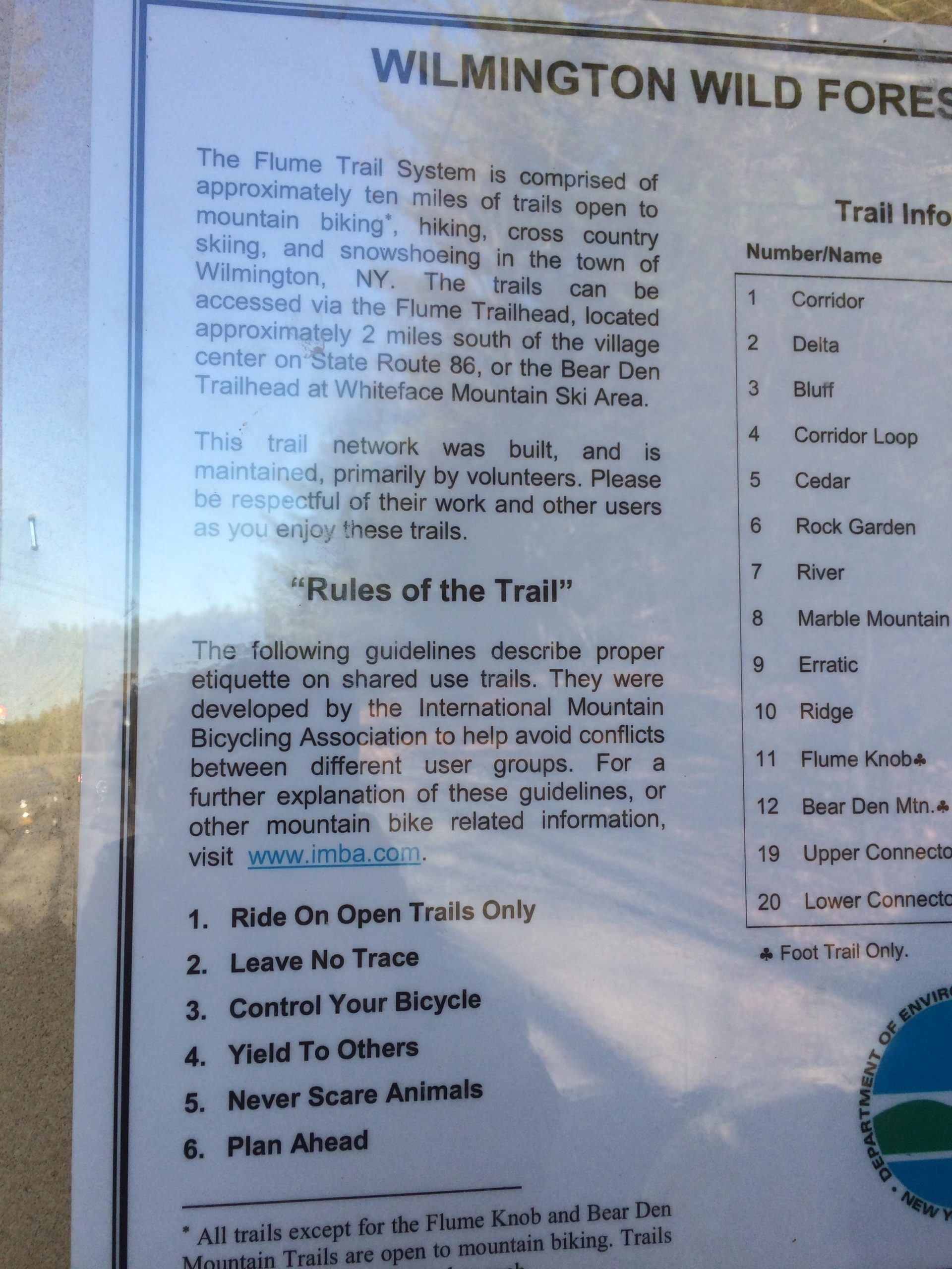 Signage for the Flume Trail System in Wilmington, NY, detailing guidelines for mountain biking, hiking, and other outdoor activities. The sign includes a description of the trail network and lists rules for sharing the trails, such as "Ride On Open Trails Only" and "Leave No Trace." The sign also includes a list of trail names and their corresponding numbers. Flume Trails mountain bike trail.