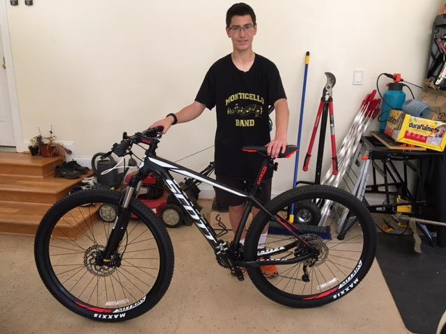 Scott Scale 970: A young person stands next to a mountain bike in a workshop setting. The bike is black and red with large tires, and the person is wearing a black t-shirt with "Monticello Band" printed on it. Various tools and equipment are visible in the background.