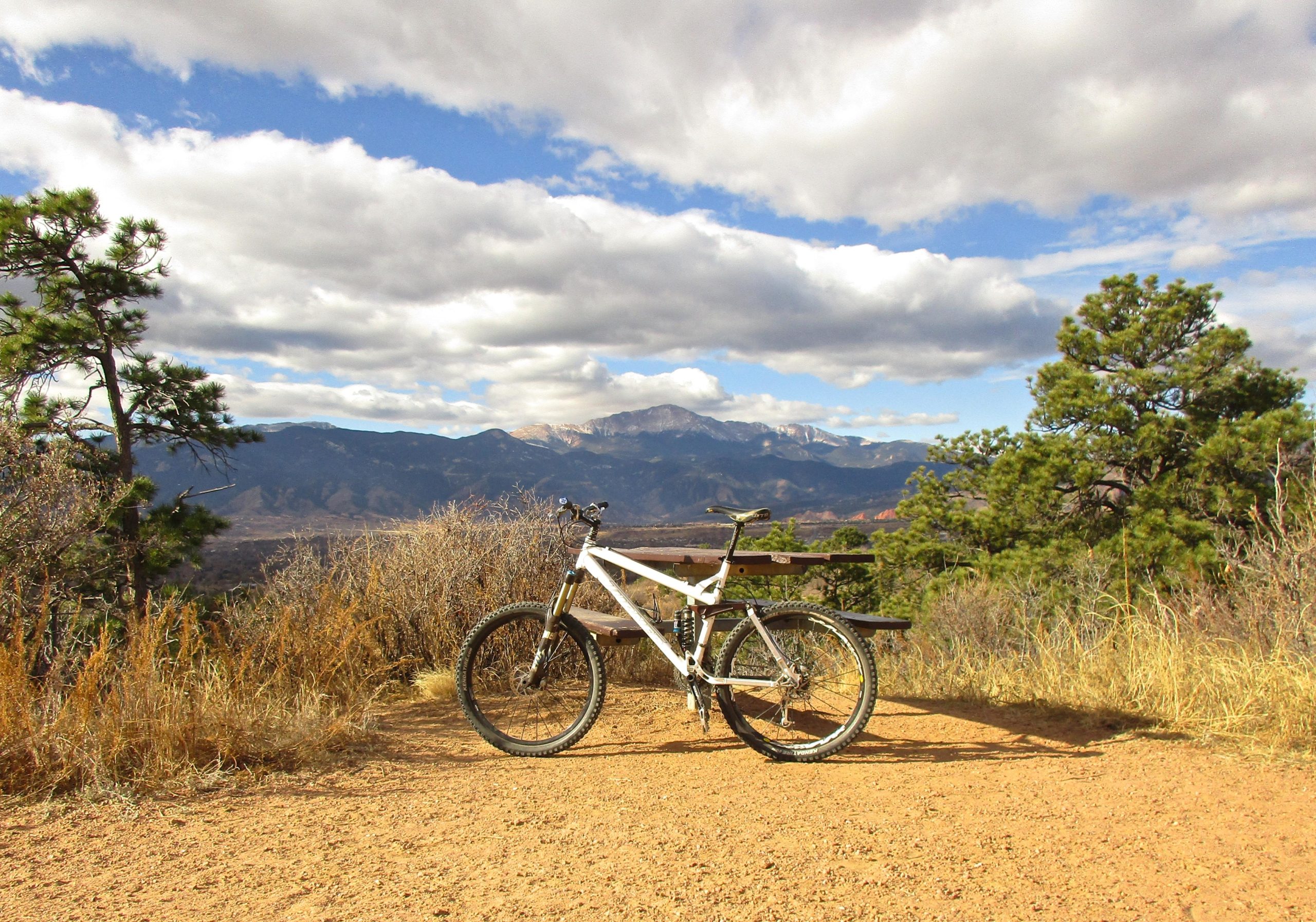 A mountain bike parked on a dirt trail overlooking a scenic mountain landscape, with a few clouds in the sky and pine trees in the foreground. Palmer Park mountain bike trail.