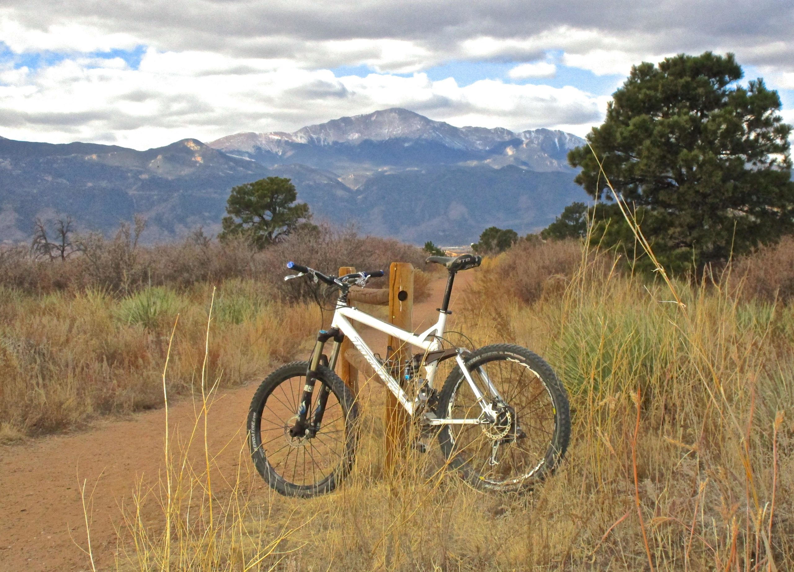 A white mountain bike is parked next to a wooden post along a dirt trail, surrounded by tall grass and shrubs. In the background, rolling mountains are visible under a cloudy sky. The scene conveys a sense of outdoor adventure and natural beauty. Palmer Park mountain bike trail.