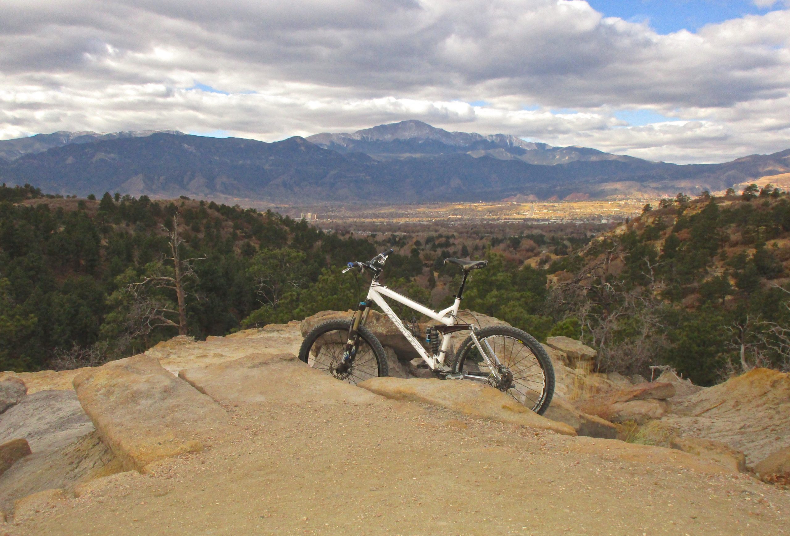 A mountain bike resting on a rocky outcrop, with a panoramic view of mountains and a valley in the background under a cloudy sky. Palmer Park mountain bike trail.