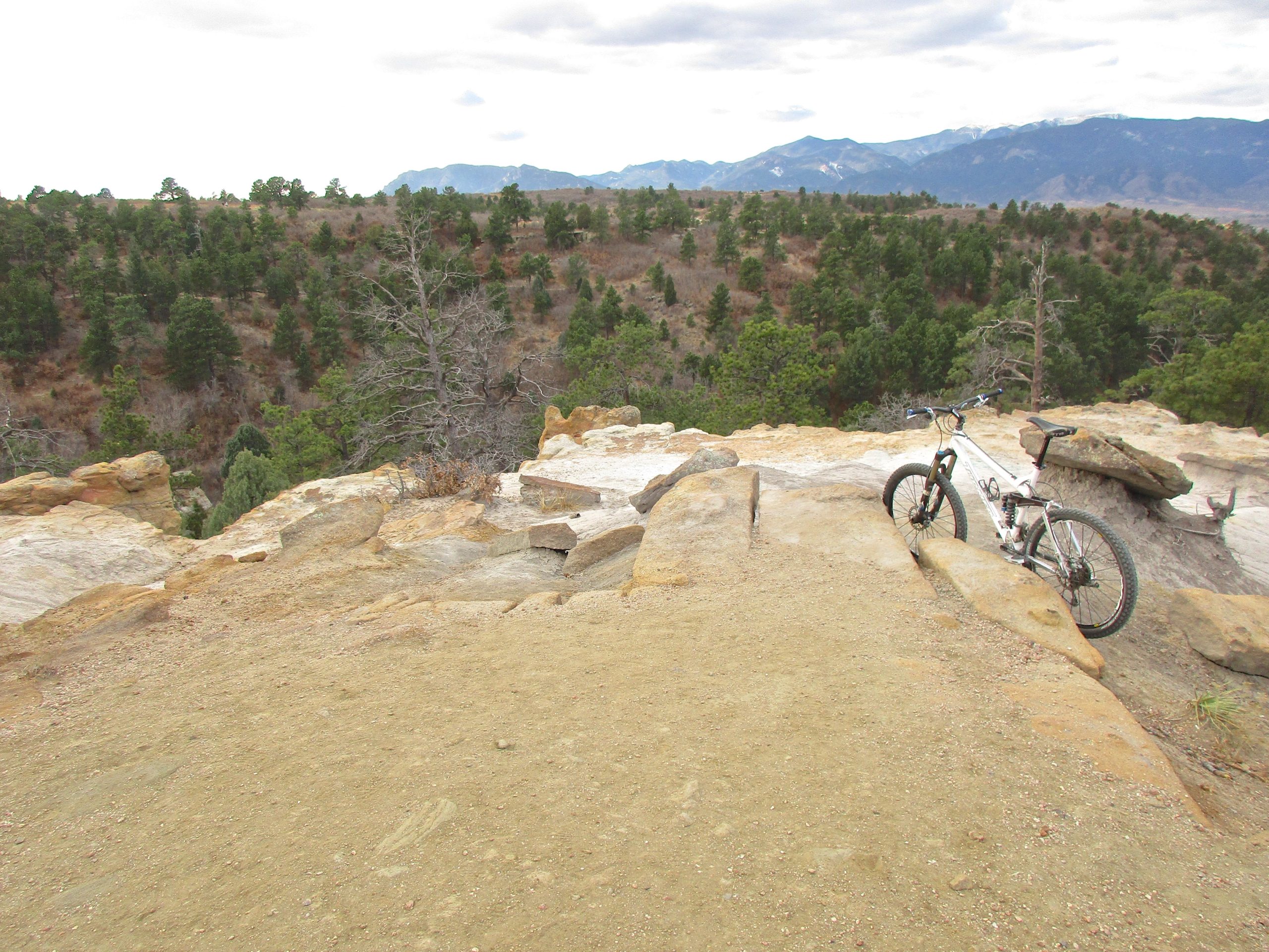 A mountain bike resting on rocky terrain overlooking a lush valley with scattered trees and distant mountains under a partly cloudy sky. Palmer Park mountain bike trail.