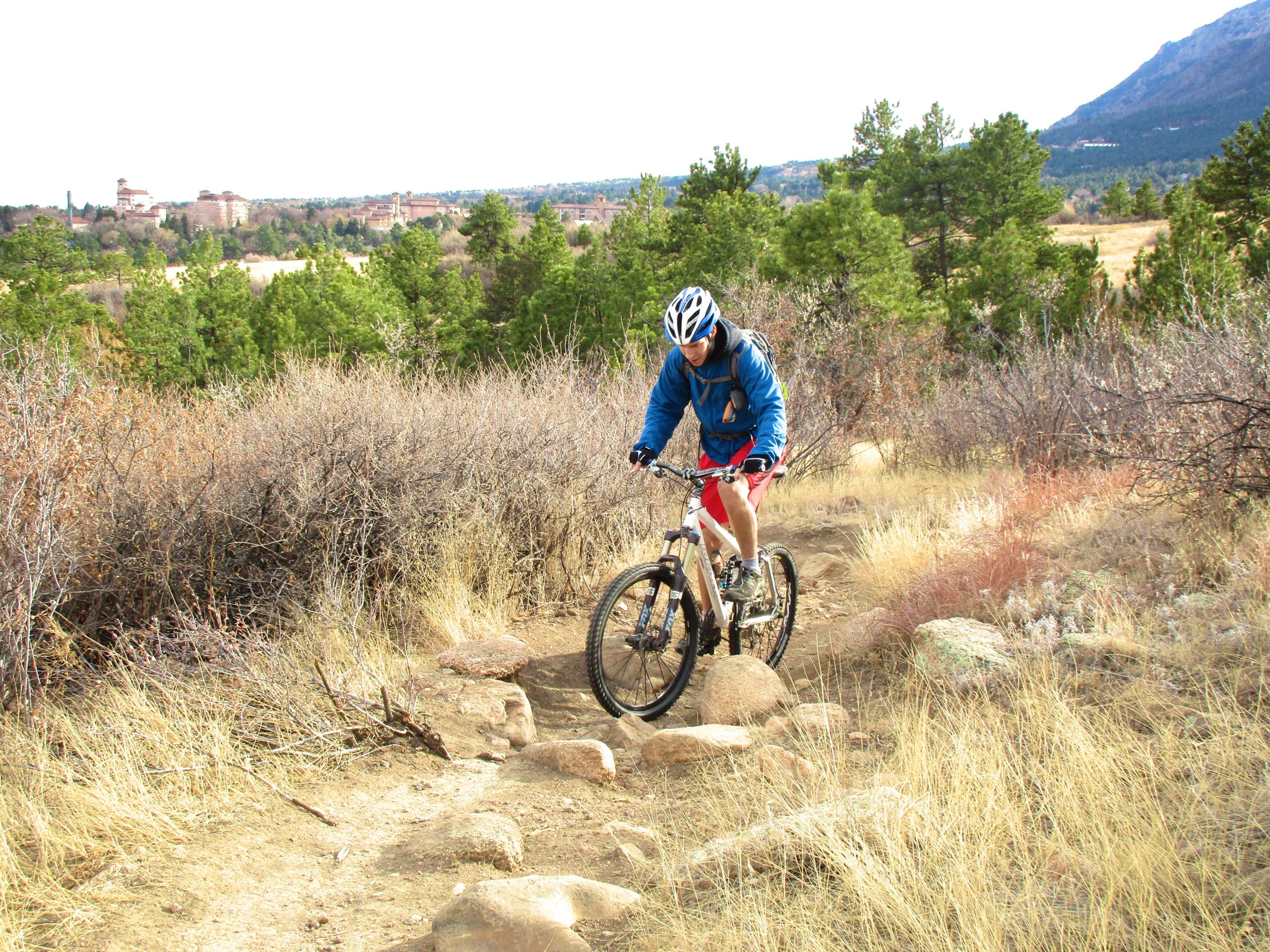 A person riding a mountain bike on a rocky trail surrounded by sparse vegetation and trees, with a mountainous background and a distant view of buildings. The cyclist is wearing a blue jacket and a helmet, focused on navigating the terrain. Stratton Open Space / The Chutes mountain bike trail.