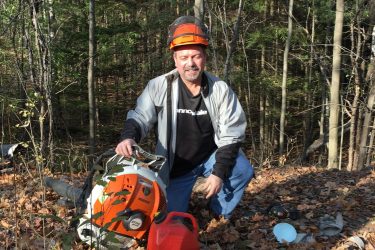 A person kneeling in a wooded area covered with fallen leaves, holding a gas canister next to a leaf blower. They are wearing a safety helmet and a light jacket, surrounded by trees in the background. Dagmar North mountain bike trail.