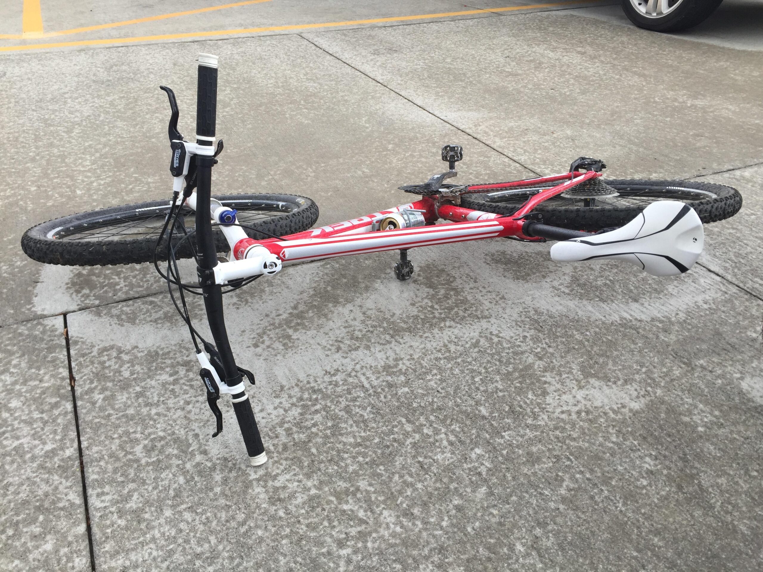 Trek Cobia: A red and white mountain bike lies on its side on a concrete surface, with one wheel partially submerged in a puddle. The handlebars and seat are visible, and the bike appears to be in a resting position, suggesting it may have fallen over.