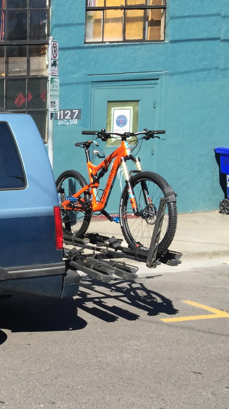 Lapierre Zesty AM 427: A bright orange mountain bike secured on a bike rack attached to the back of a blue vehicle, parked in front of a teal building with large windows and a door labeled "suite 100." A nearby street sign indicates no parking restrictions.