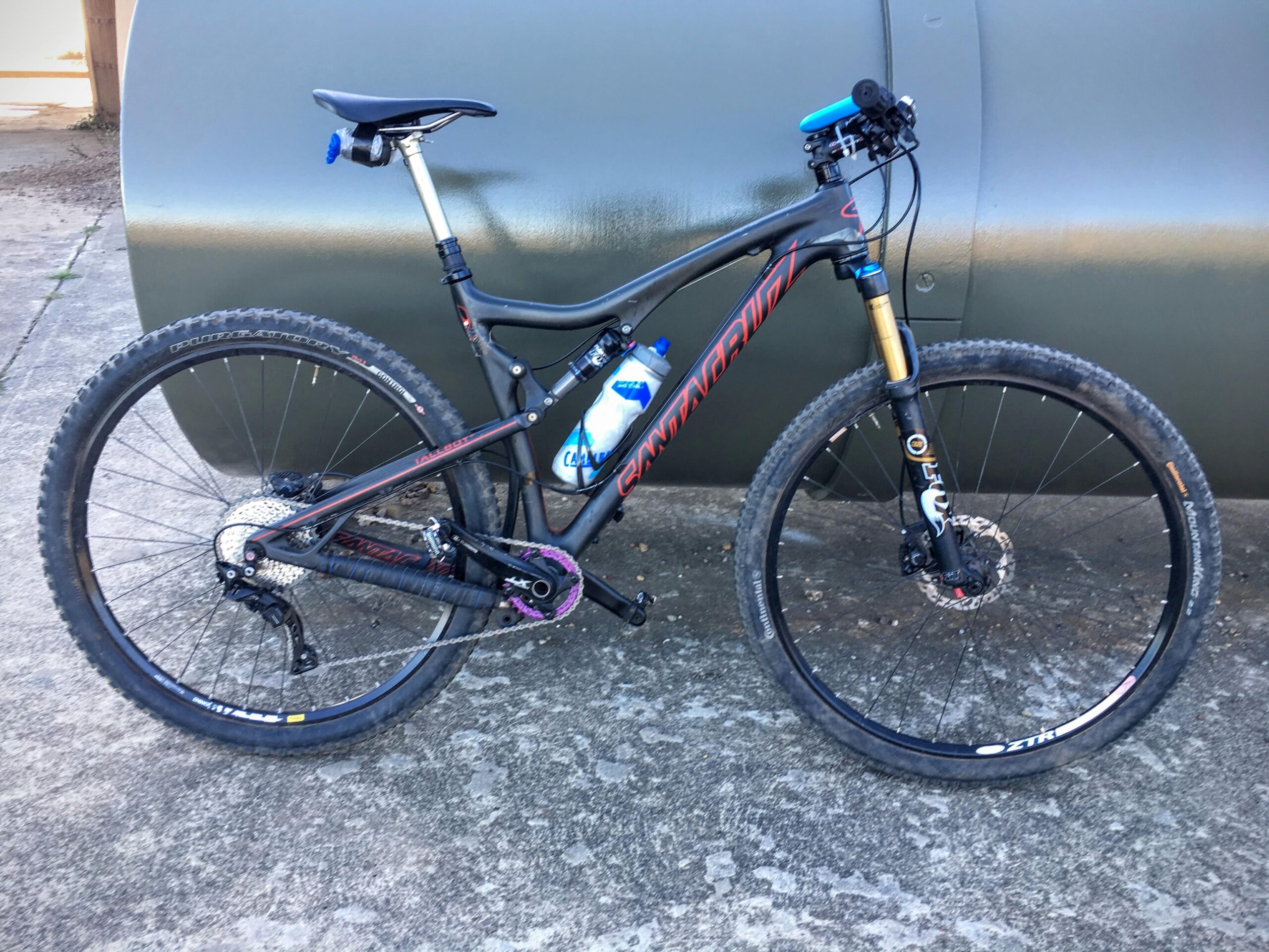 Santa Cruz Tallboy Carbon: A mountain bike with a black frame, featuring red and blue accents, stands against a gray cylindrical object. The bike has thick tires suitable for off-road conditions, a water bottle mounted on the frame, and a prominent front suspension fork. The background shows a concrete surface with some gravel.