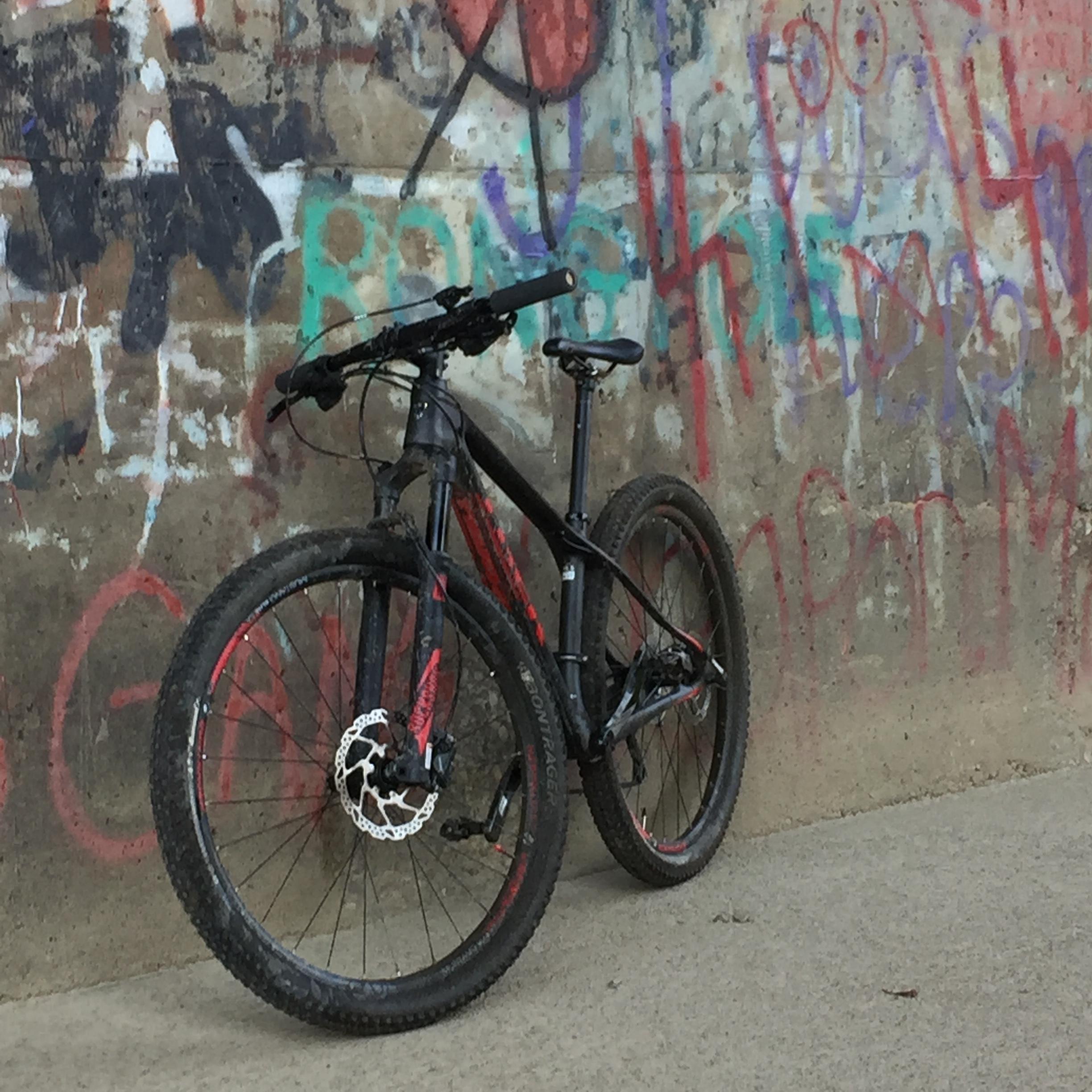 A black mountain bike with red accents leans against a graffiti-covered concrete wall. The bike shows signs of use, with mud on the tires and frame. Shenango River Trail mountain bike trail.