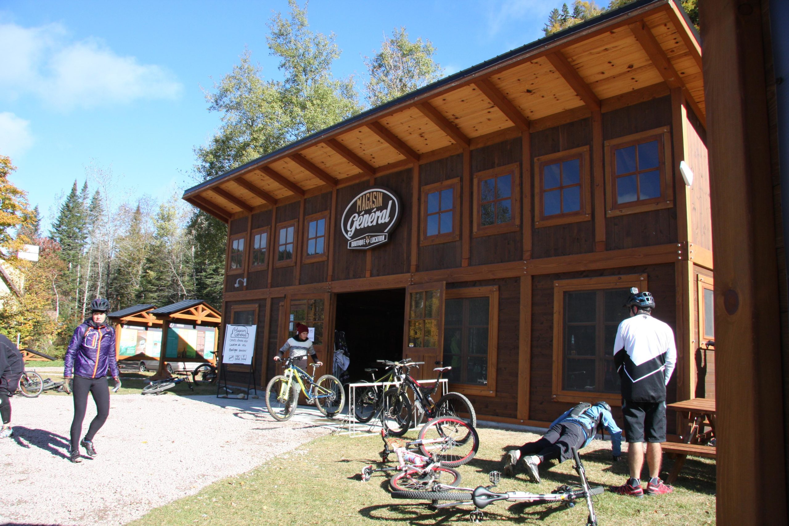 A rustic wooden general store with a sign, surrounded by trees on a clear day. Several bicycles are parked outside, and two people are interacting in front of the store, while another person is doing push-ups nearby. The scene conveys an active outdoor atmosphere. Vallee Bras Du Nord Secteur Shannahan mountain bike trail.