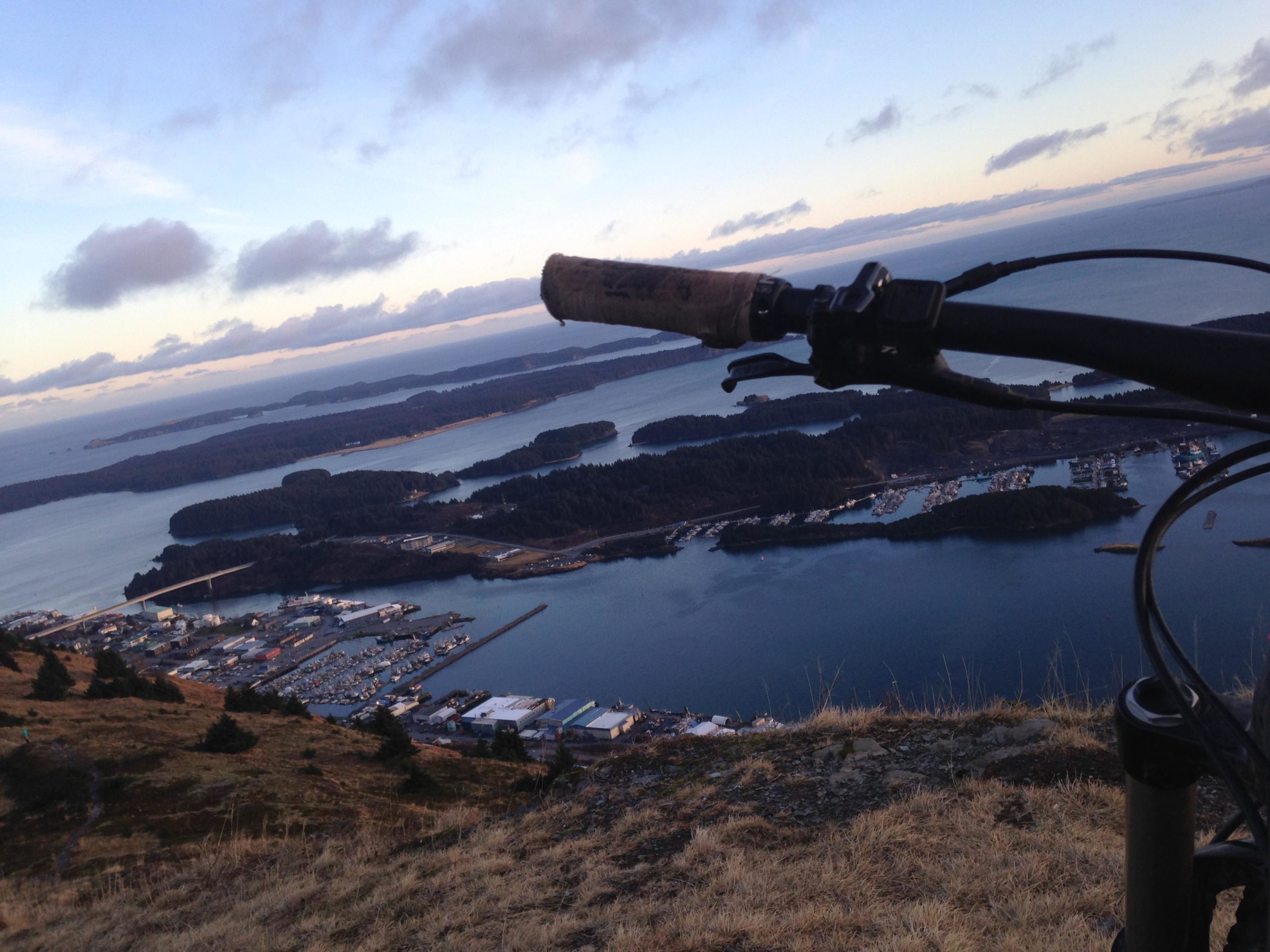 A scenic view from a hillside overlooking a marina and surrounding islands, with the handlebars of a mountain bike in the foreground. The sky is partly cloudy, and the landscape features a mix of water, land, and boats in the harbor below. Pillar mountain bike trail.