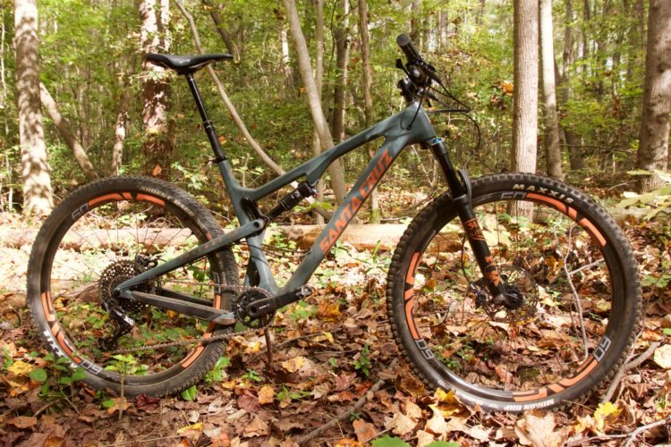 A mountain bike with a sleek design and orange accents positioned on a forest floor covered in autumn leaves. The bike features thick tires suitable for rugged terrain, a black and gray frame, and is surrounded by trees in a lush green environment.