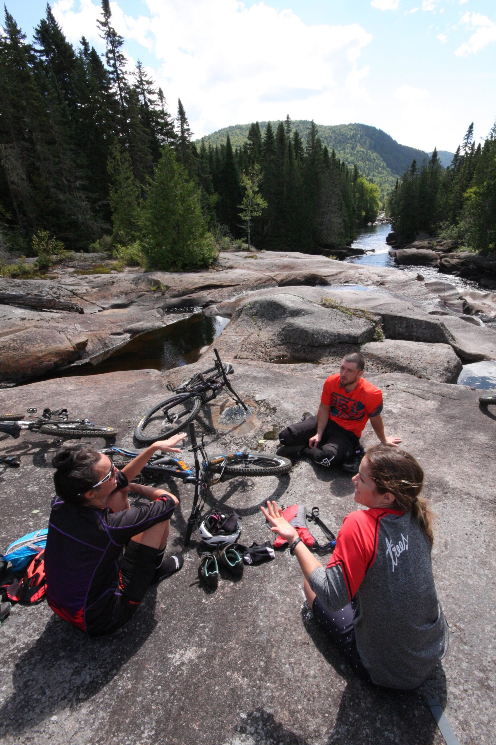 Three mountain bikers are sitting on rocky terrain near a stream, surrounded by trees and hills in the background. Two people are engaged in conversation while the third listens, and their mountain bikes are propped up nearby. Personal gear, including shoes and clothing, is scattered around them. The scene captures a moment of relaxation in a natural setting. Vallee Bras Du Nord Secteur Shannahan mountain bike trail.