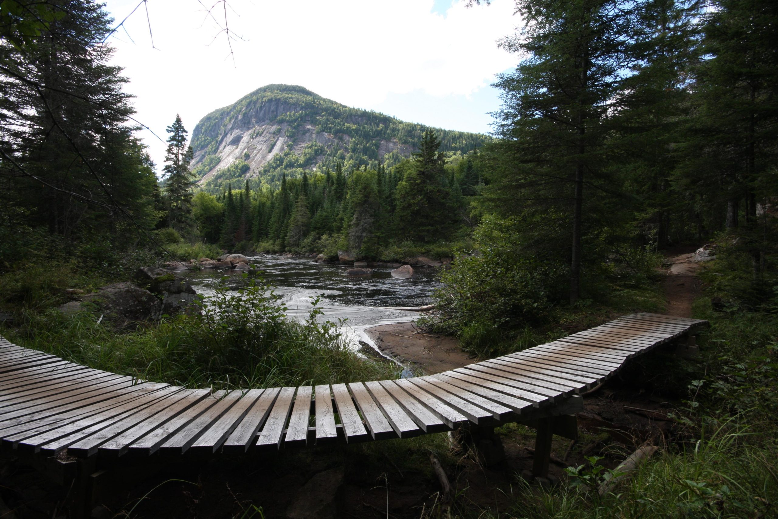 A wooden boardwalk curves along the edge of a river, surrounded by lush greenery and dense trees. In the background, a rugged mountain rises, partially obscured by clouds, creating a serene natural landscape. Vallee Bras Du Nord Secteur Shannahan mountain bike trail.