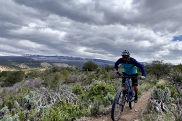 A cyclist wearing a blue and black outfit rides a mountain bike along a narrow dirt trail surrounded by shrubs and trees, with mountains covered in snow visible in the background under a cloudy sky. Ridgway Area Trails mountain bike trail.