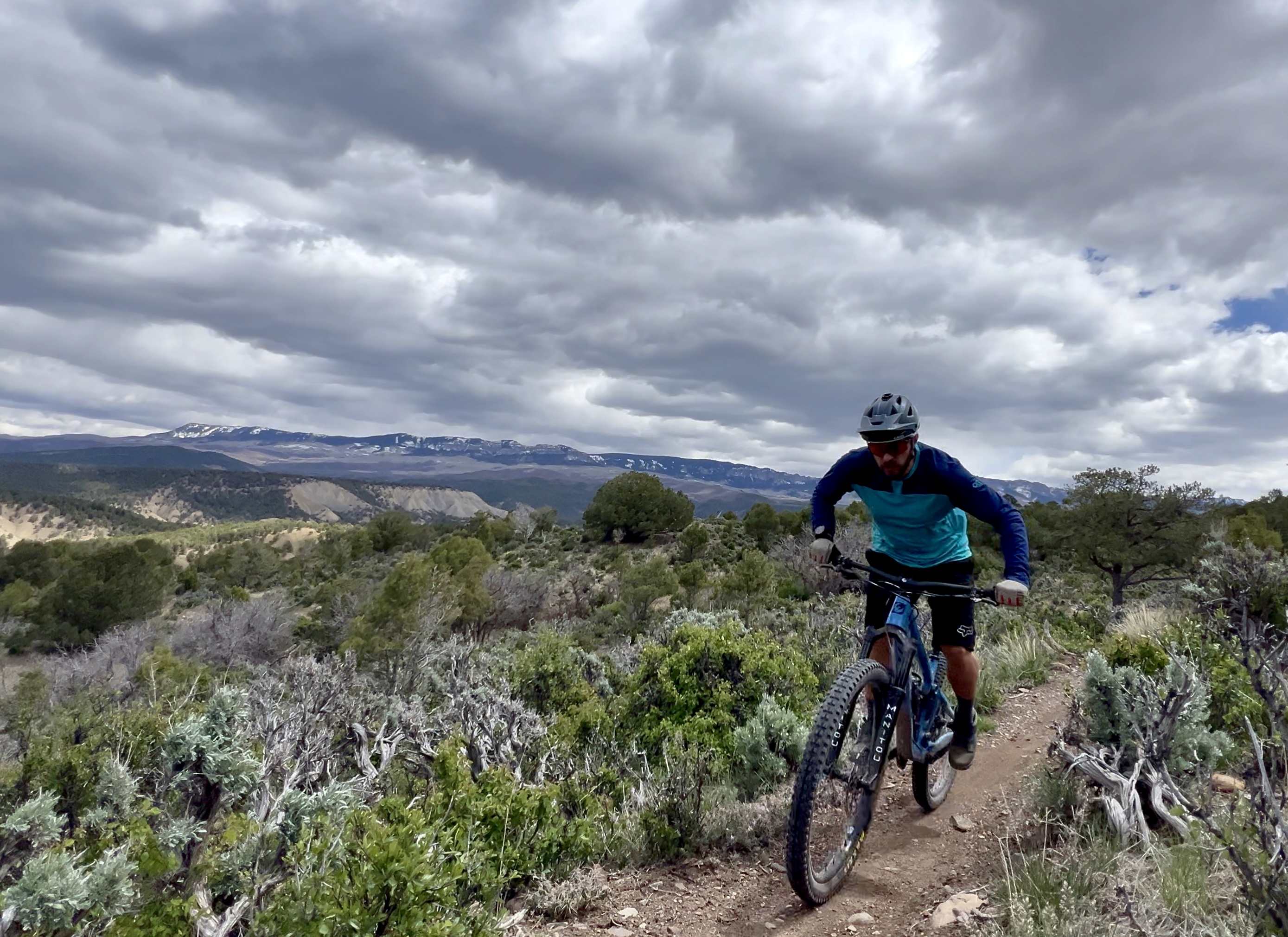 A cyclist wearing a blue and black outfit rides a mountain bike along a narrow dirt trail surrounded by shrubs and trees, with mountains covered in snow visible in the background under a cloudy sky. Ridgway Area Trails mountain bike trail.