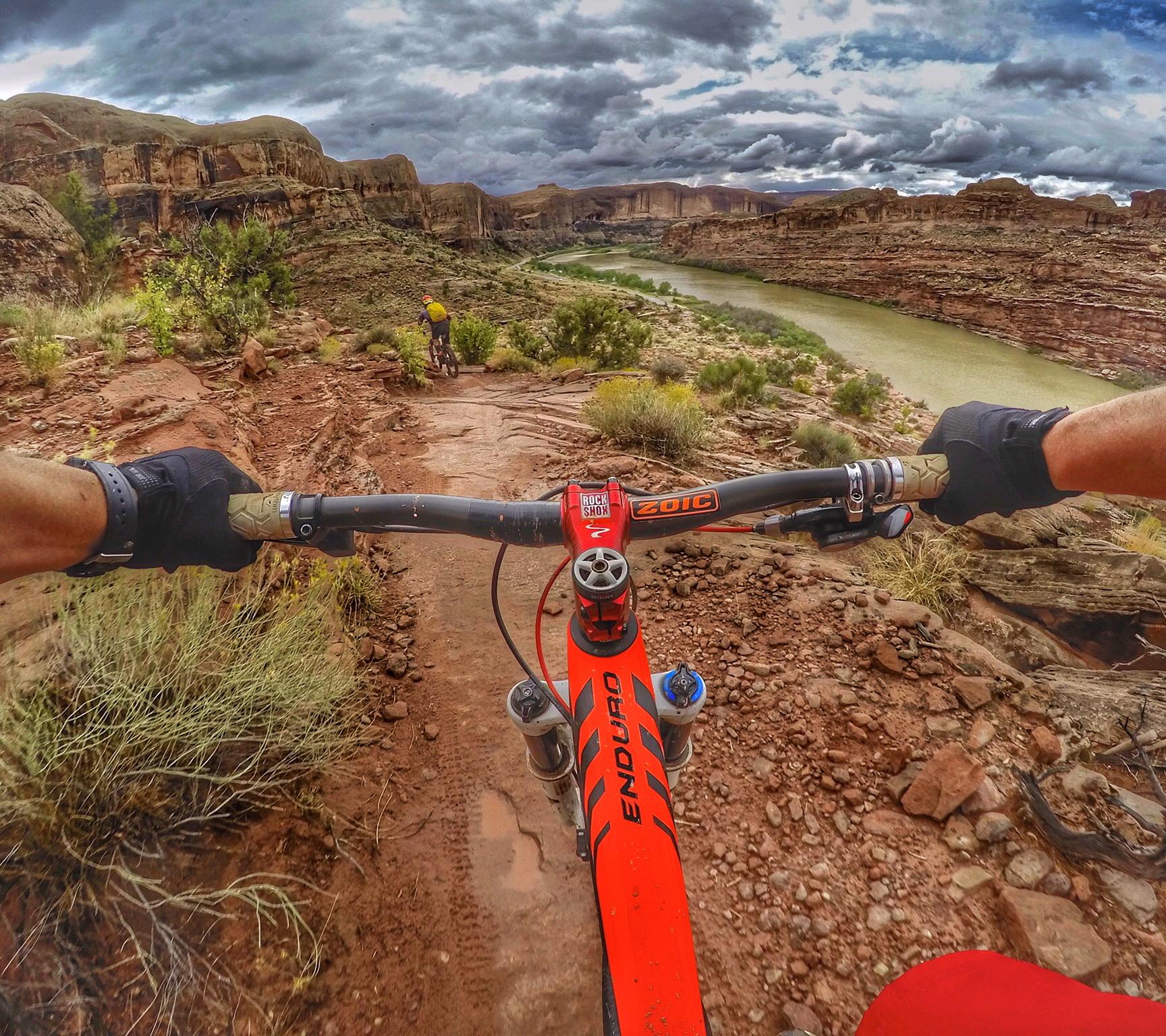 A mountain biker's view from the handlebars on a rocky trail, overlooking a river and rugged landscape under a dramatic sky filled with clouds. The bike features an orange frame and the rider is navigating a downhill path with another cyclist visible in the background. Porcupine Rim mountain bike trail.