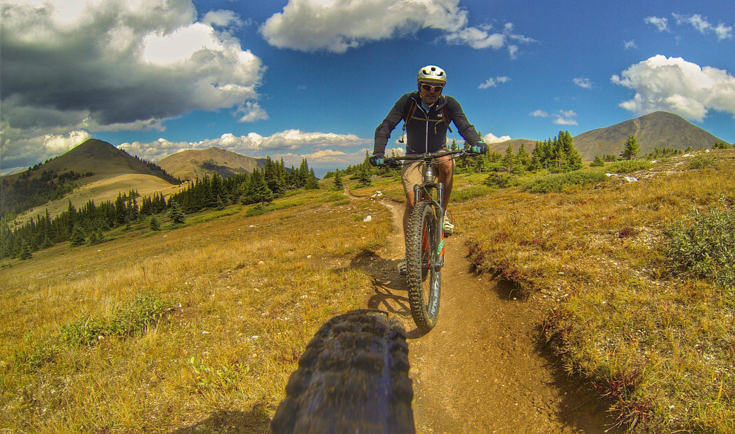 A person riding a mountain bike on a dirt trail in a scenic mountainous area, with a backdrop of hills, trees, and a partly cloudy blue sky. The cyclist is wearing a helmet and sports gear, enjoying the outdoor adventure. Monarch Crest Trail mountain bike trail.