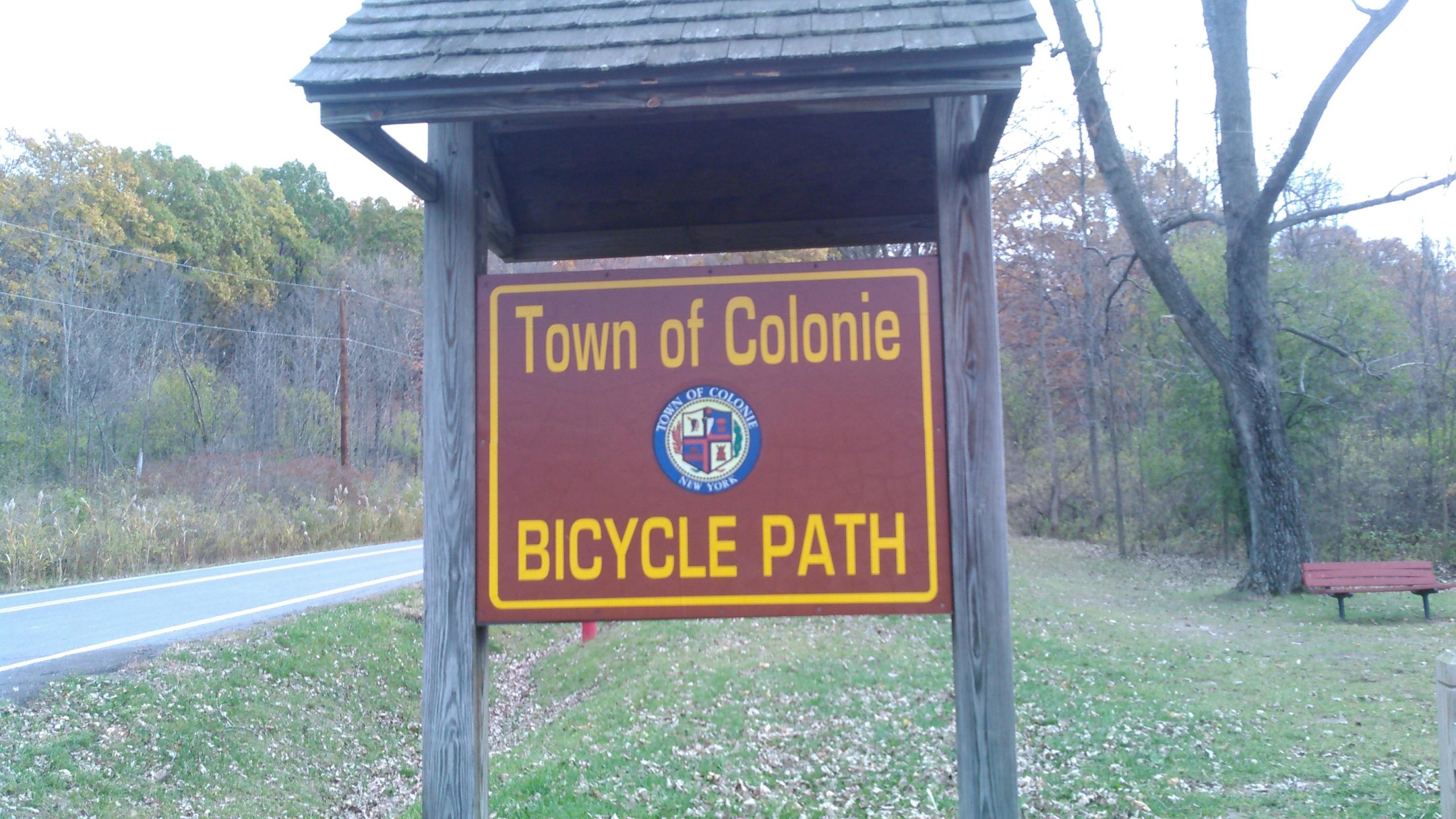 Sign indicating the "Town of Colonie Bicycle Path," featuring the town emblem and surrounded by trees and grass, with a road visible in the background. Colonie Town Park/Ridge Trail mountain bike trail.