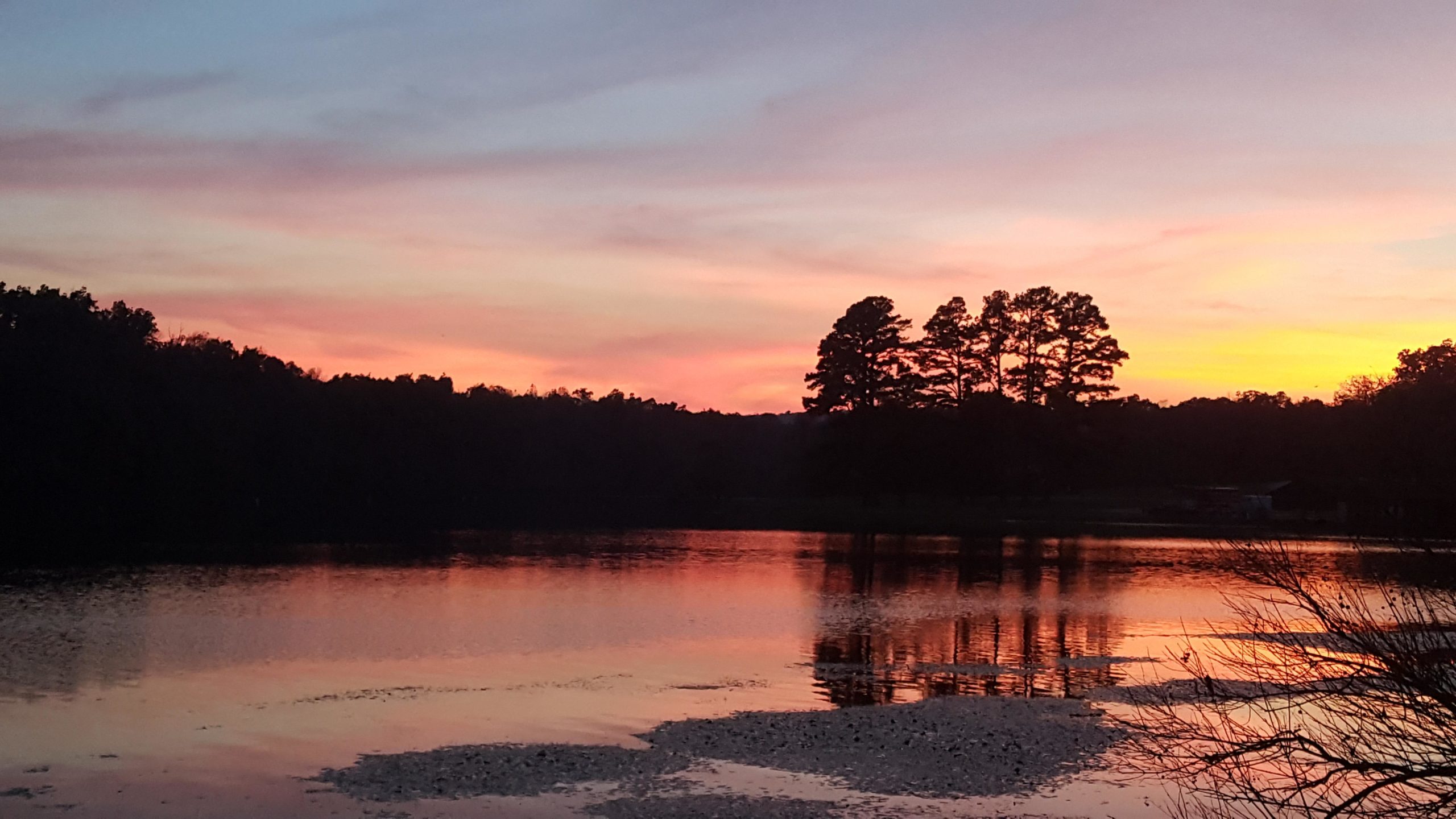 A serene lakeside view at sunset, showcasing vibrant hues of pink, orange, and blue in the sky. The calm water reflects the colors and silhouettes of distant trees, creating a peaceful atmosphere. Lake Wedington Trail mountain bike trail.