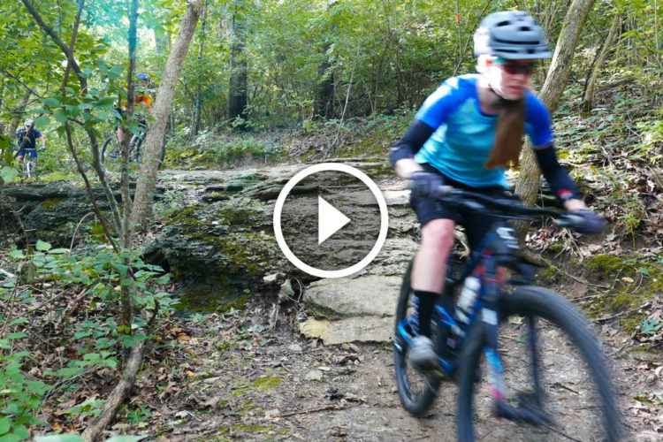 A mountain biker rides along a rocky trail surrounded by lush greenery in a forested area. The rider, wearing a helmet and a blue shirt, is in motion, suggesting an active, adventurous atmosphere. Additional bikers can be seen in the background, contributing to the outdoor cycling experience. A play button indicates that the image is part of a video.