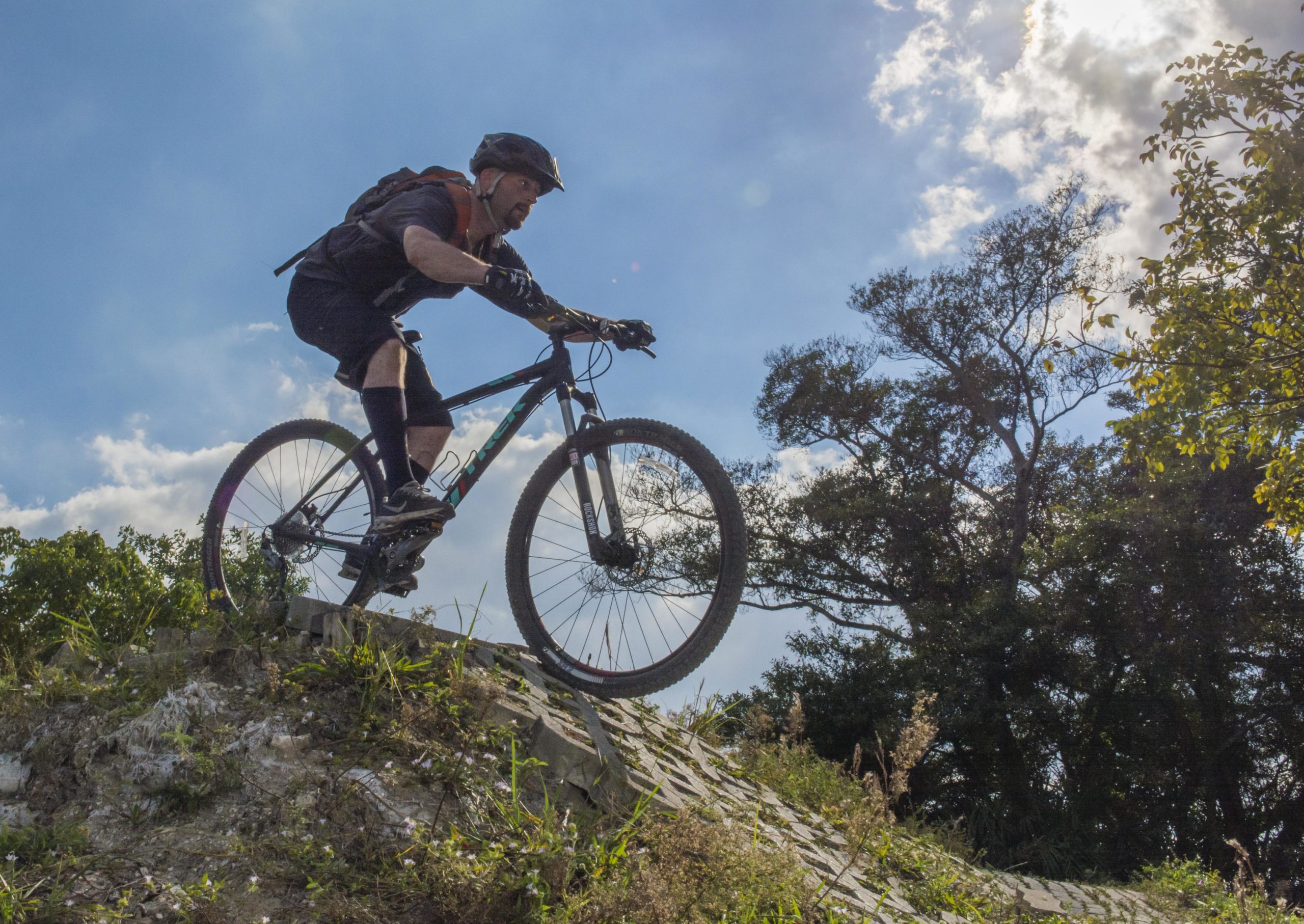 A mountain biker riding on a steep slope, airborne as he descends, with a clear blue sky and scattered clouds in the background. The scene is surrounded by greenery and trees. Quiet Waters Park mountain bike trail.