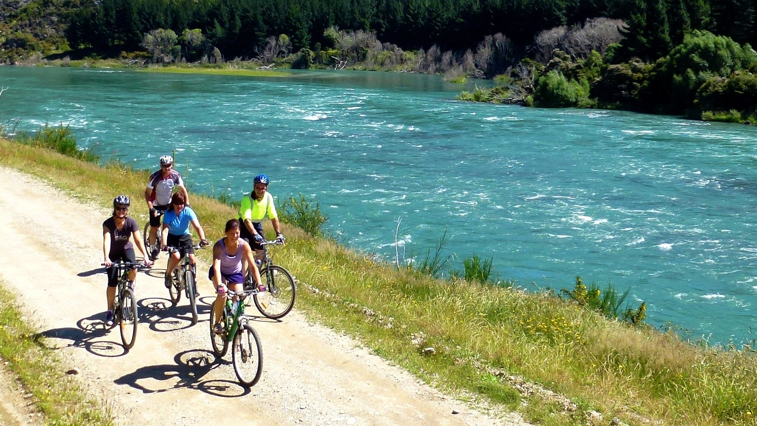 A group of five people riding bicycles along a dirt path next to a vibrant turquoise river, surrounded by greenery and trees. The scene is set in a sunny environment, showcasing an outdoor recreational activity. Clutha Gold Trail mountain bike trail.