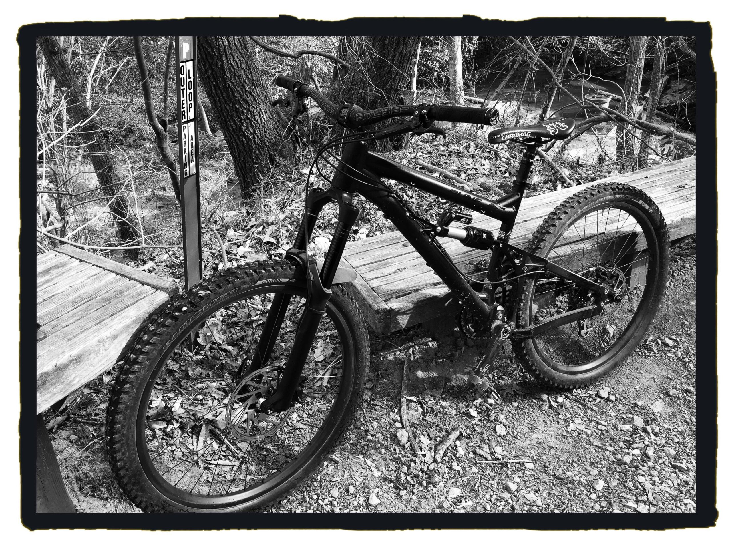 Banshee Rune: A black and white image of a mountain bike resting on a wooden platform by a wooded area. The bike features a sturdy frame, thick tires, and a front suspension. In the background, there's a sign indicating parking information, with trees and foliage surrounding the scene.