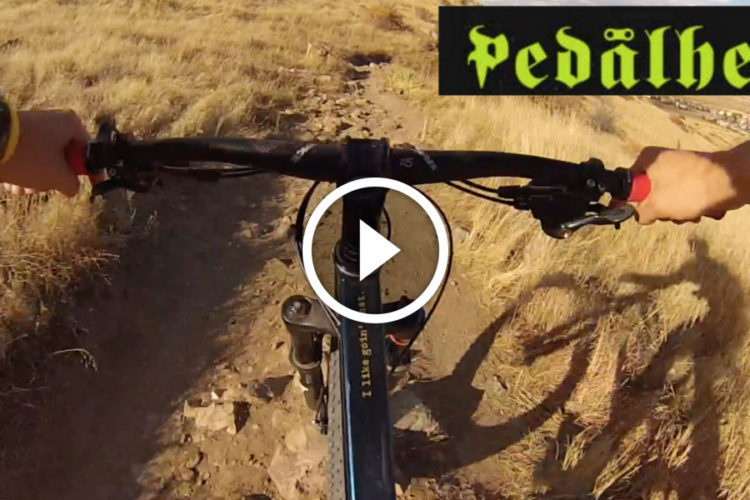 A close-up view of handlebars on a mountain bike, with a hand gripping the right handlebar brake. The background shows a rocky dirt trail surrounded by dry grass. The top left corner features the text "Pedalhead" in a stylized font. A play button icon is positioned in the center of the image, suggesting video content.