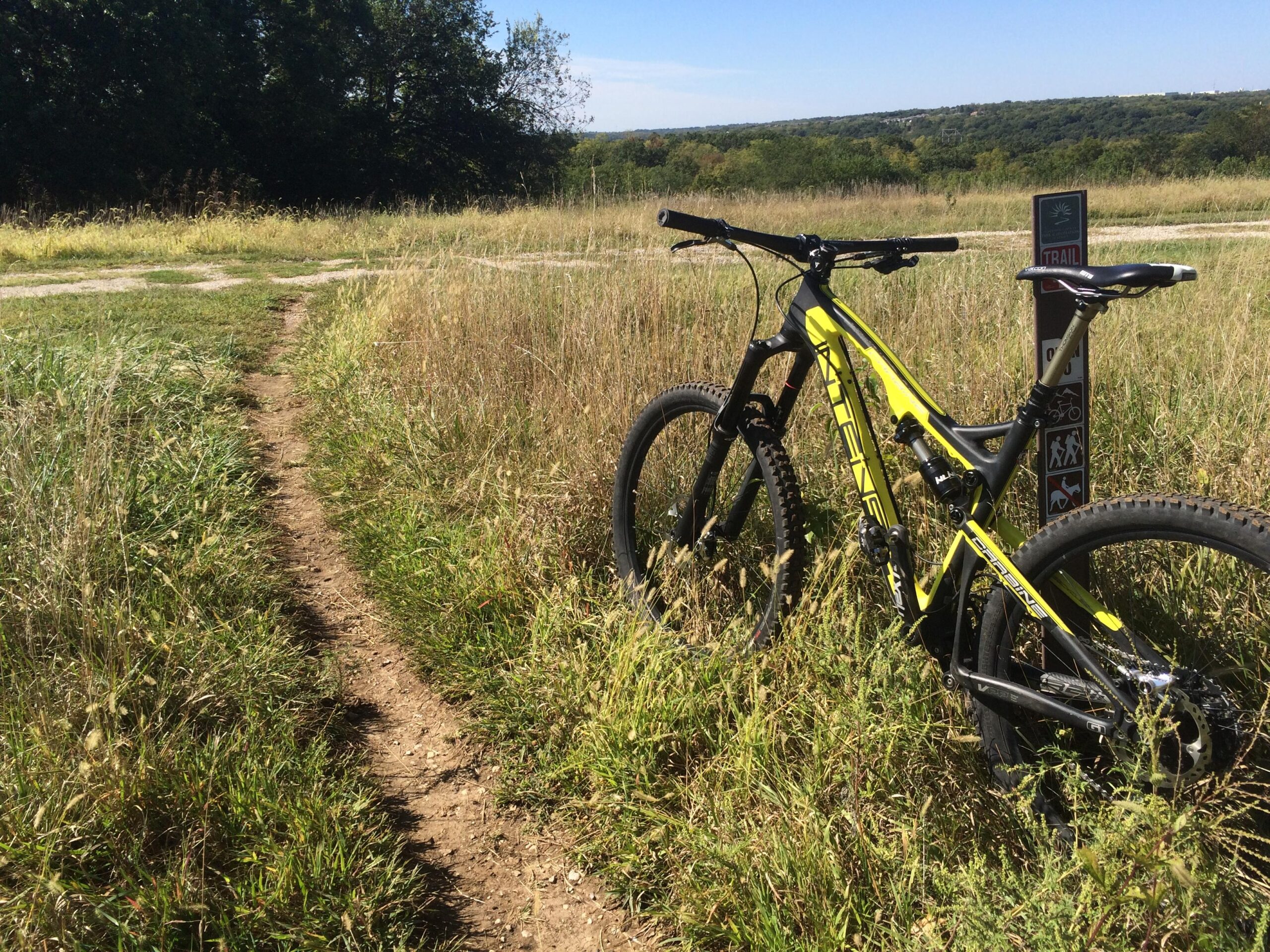 Intense Carbine SL: A bright yellow mountain bike is leaning against a trail sign in a grassy field. A dirt path leads away from the bike, winding through the tall grass under a clear blue sky. The scene features a serene natural landscape, with trees visible in the background.