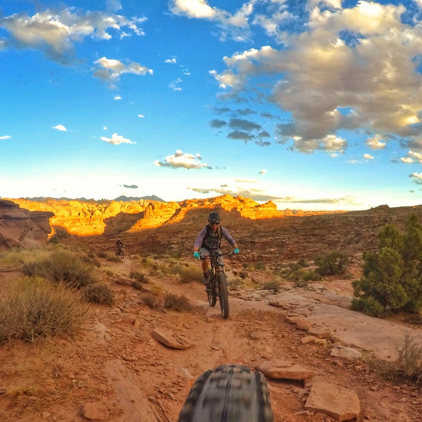 Two mountain bikers riding on a rocky trail in a desert landscape during sunset, with colorful clouds in the sky and scenic rock formations in the background. The foreground shows a close-up of a bicycle tire on the dirt path. Hymasa mountain bike trail.