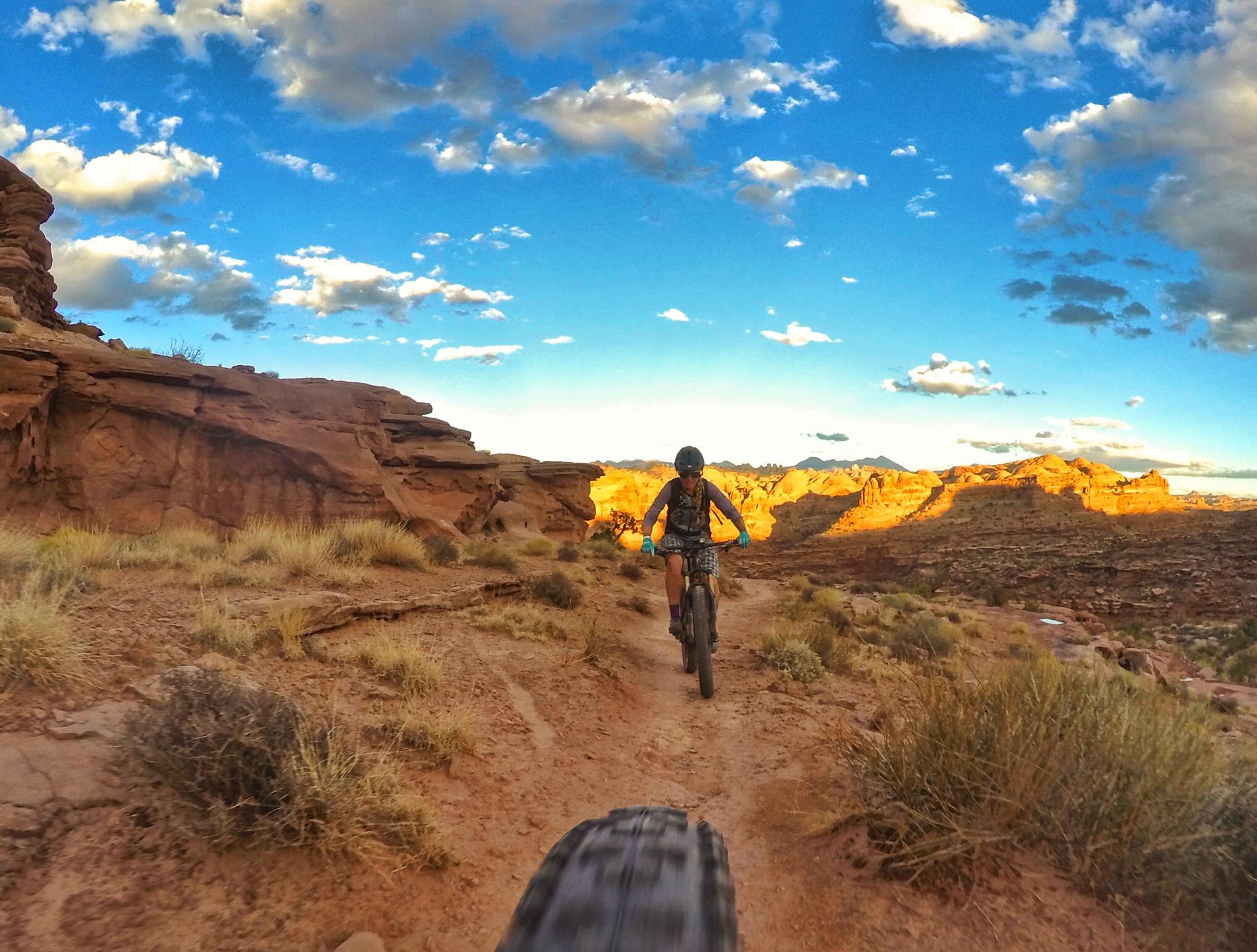 A mountain biker rides along a dirt trail surrounded by rugged, red rock formations and sparse grass. The sky is clear with wispy clouds, and the golden light of sunset illuminates the landscape in the background, creating a beautiful contrast. Hymasa mountain bike trail.