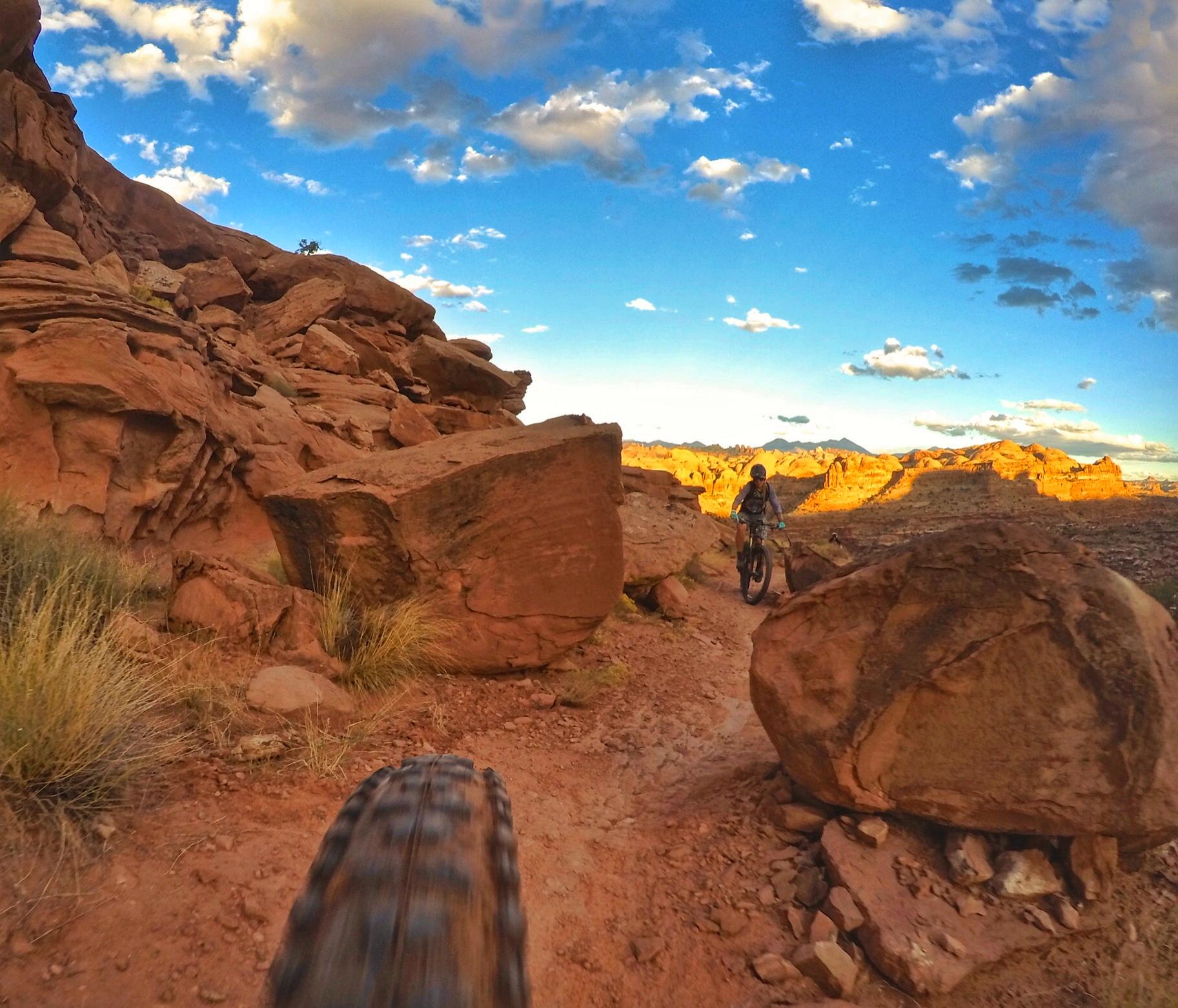 A mountain biker navigates a rocky trail surrounded by red rocks, under a blue sky with scattered clouds. The image captures the action from a low perspective, showcasing the bike's tire and the rugged terrain. Hymasa mountain bike trail.