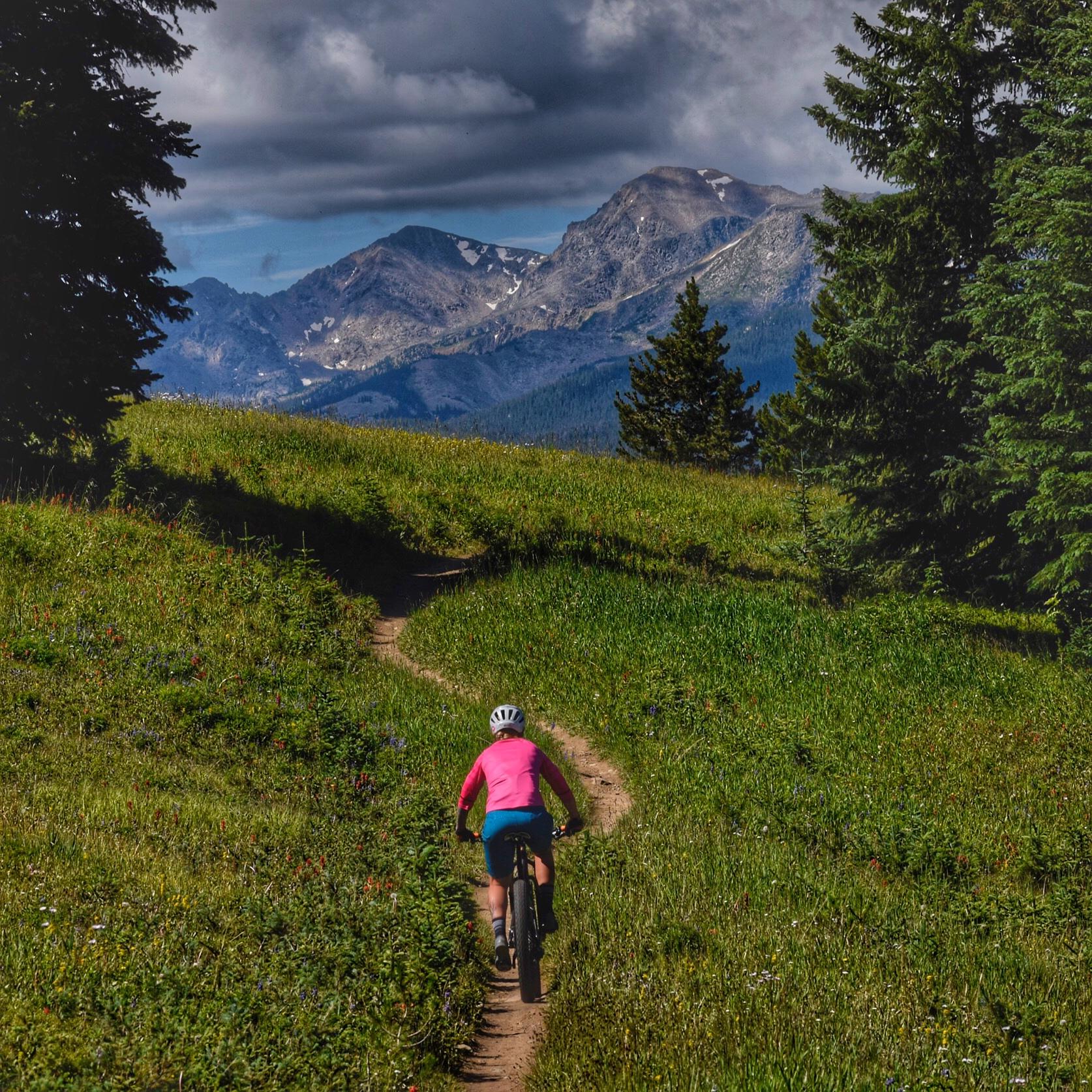A mountain biker rides along a winding dirt path through a lush green valley, with tall grass and wildflowers on either side. In the background, majestic mountains are visible under a partly cloudy sky. The cyclist is dressed in a bright pink long-sleeve shirt and blue shorts, with a white helmet on their head. Vail Mountain Bike Park mountain bike trail.