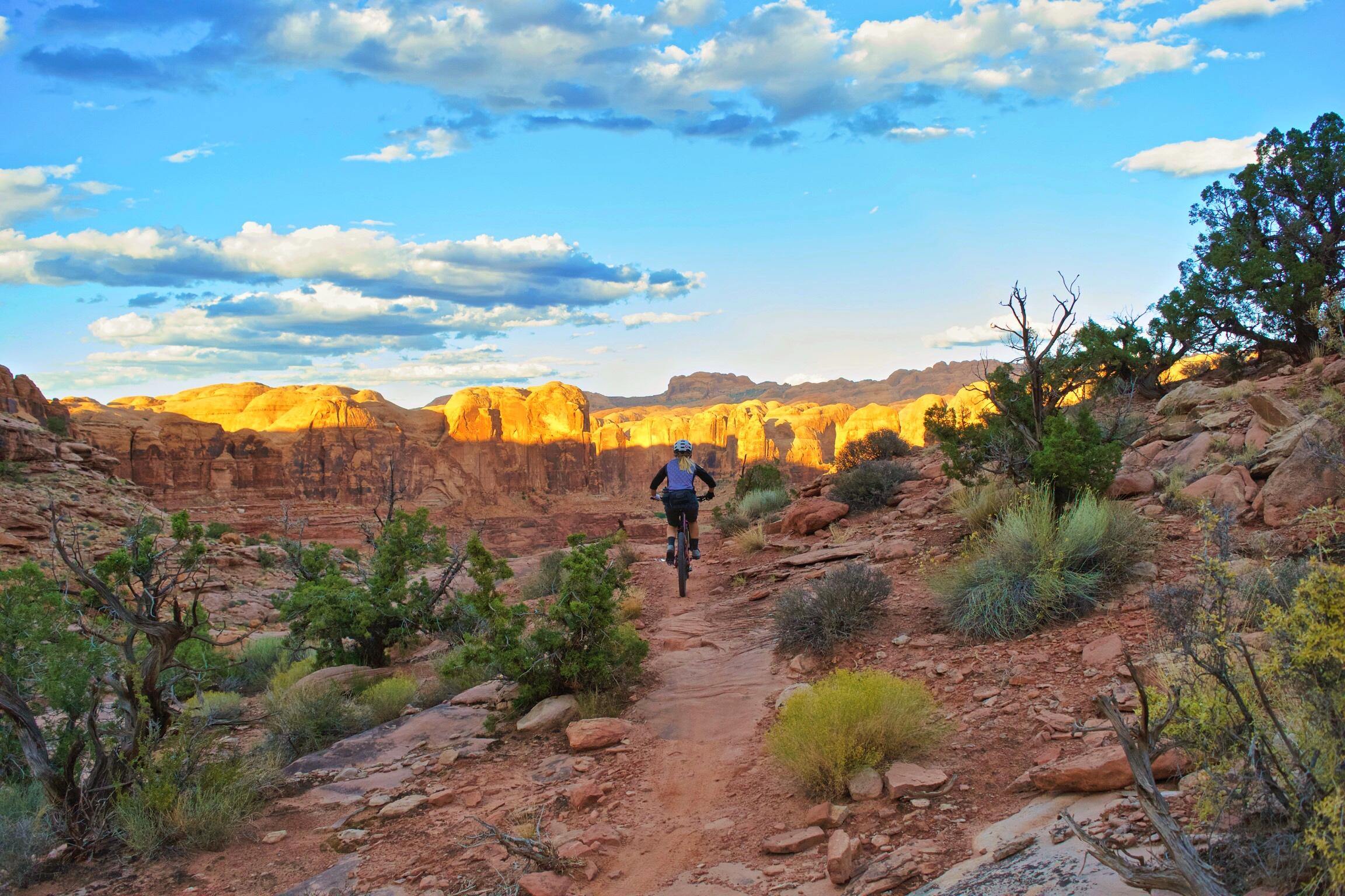 A mountain biker rides along a rocky trail in a desert landscape, surrounded by vibrant vegetation. The golden cliffs are illuminated by the setting sun, under a blue sky filled with scattered clouds. Hymasa mountain bike trail.
