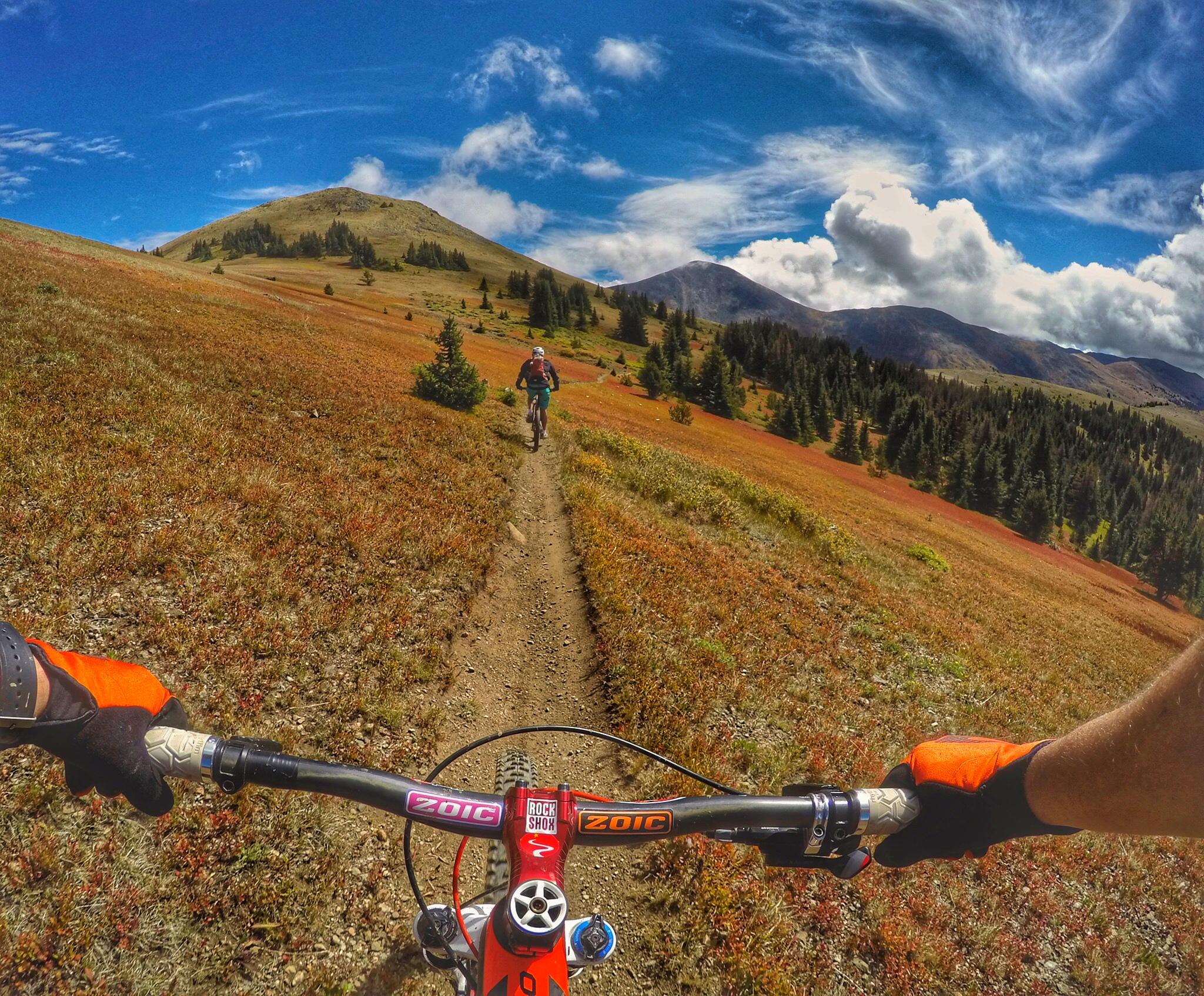 Mountain biking trail view with vibrant autumn foliage, showcasing a rider in the distance on a winding dirt path. The handlebars of the bike are in the foreground, with a clear blue sky and scattered clouds above the mountainous landscape. Monarch Crest Trail mountain bike trail.