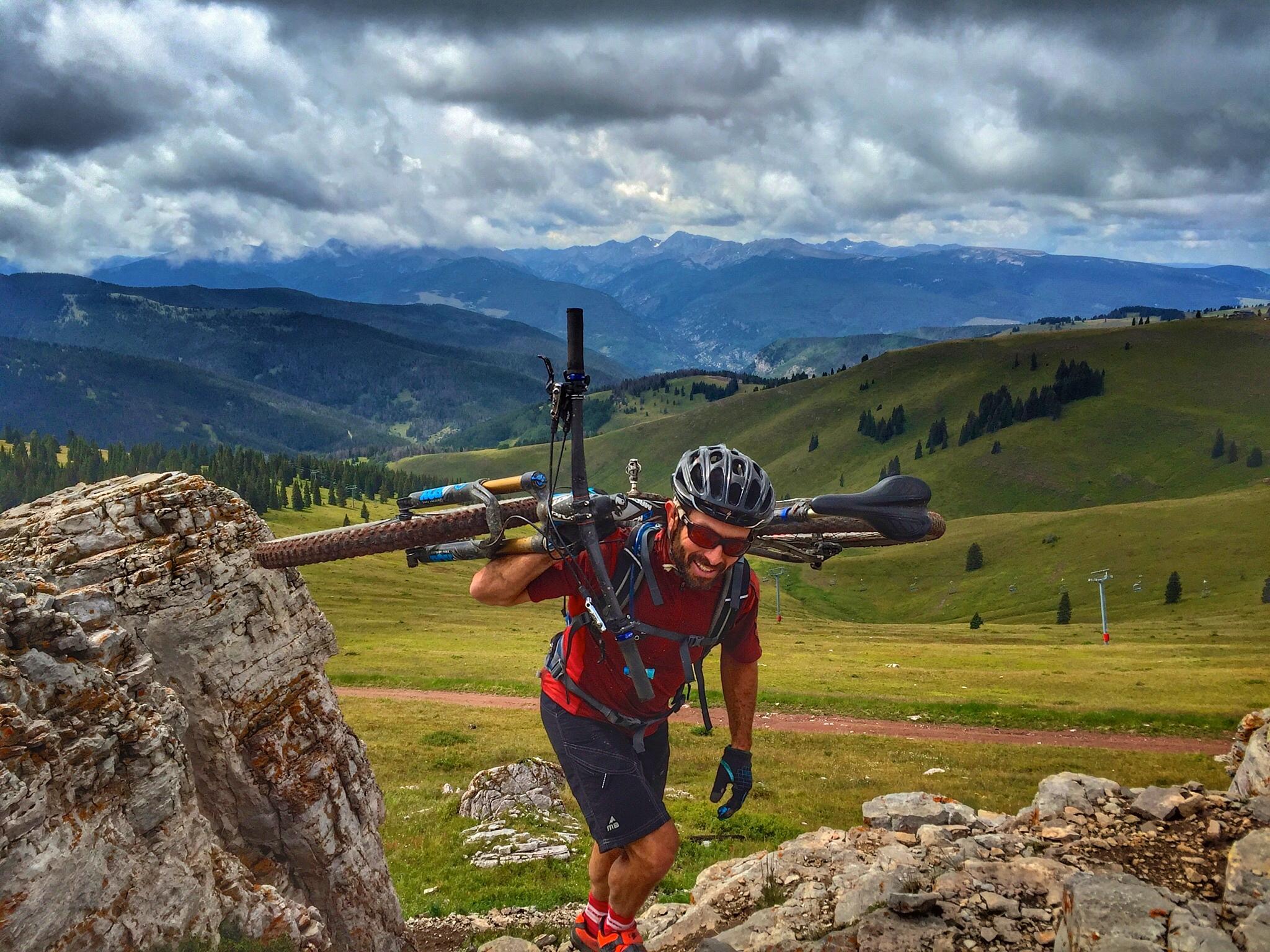 A cyclist carrying a mountain bike on their shoulder while climbing a rocky path in a mountainous landscape. The scene features lush green hills and distant mountain ranges under cloudy skies. The cyclist is wearing a helmet, gloves, and a backpack, showcasing determination as they navigate the terrain. Vail Mountain Bike Park mountain bike trail.