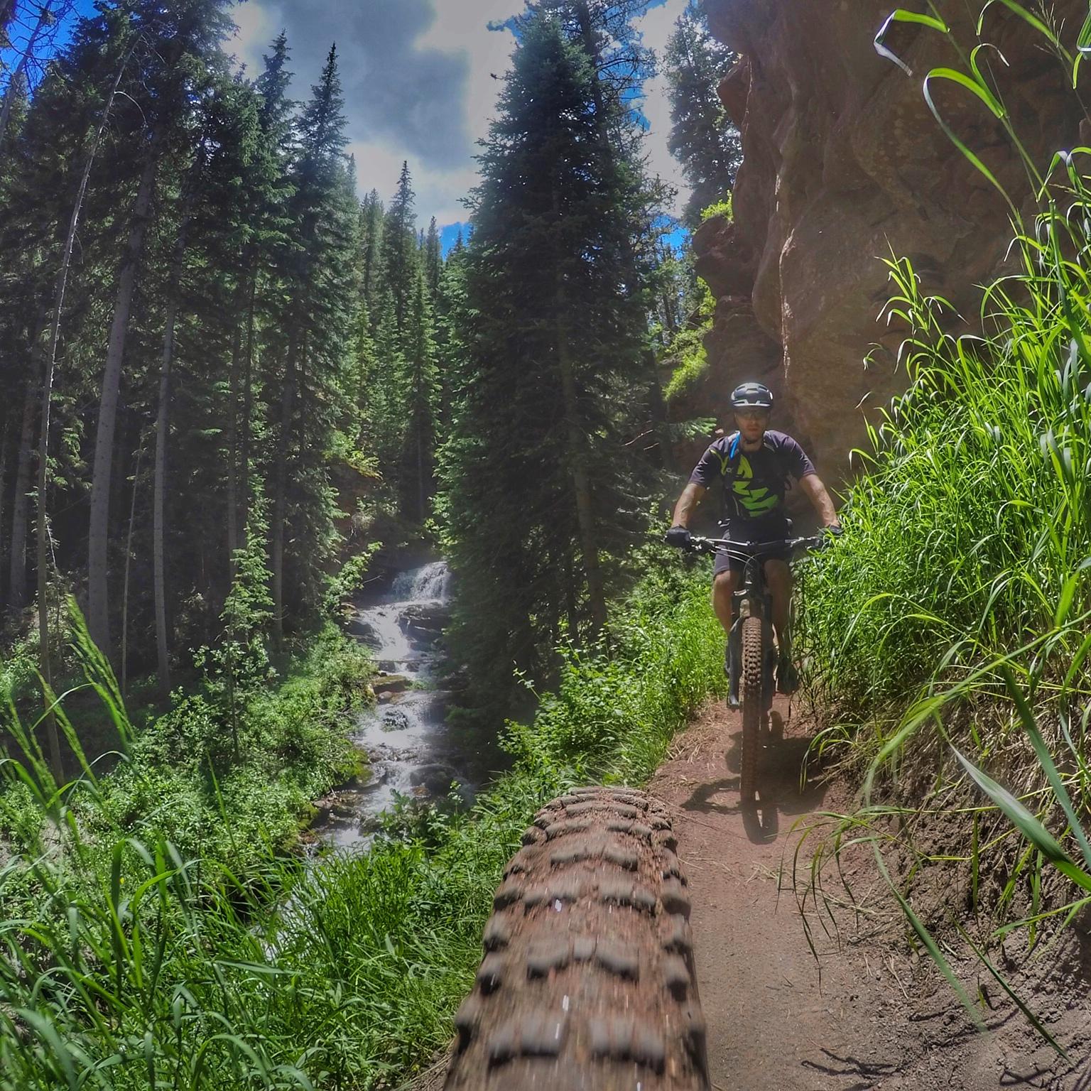 A mountain biker riding on a narrow trail surrounded by lush green trees and a flowing stream nearby, with rocky cliffs accentuating the scene under a partly cloudy sky. Vail Mountain Bike Park mountain bike trail.