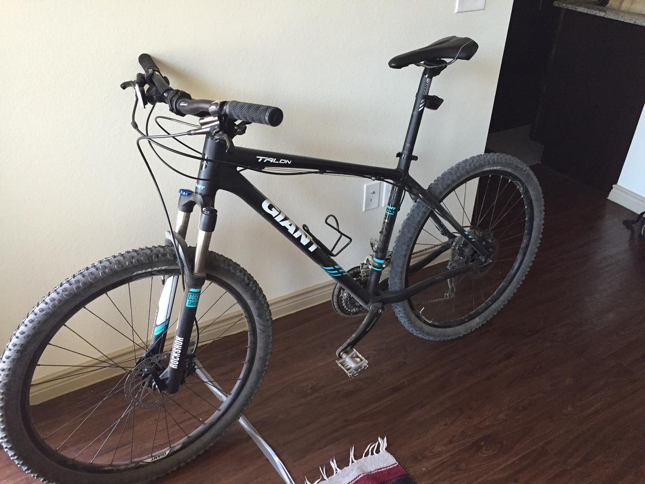 Giant Talon 27.5 2: A black mountain bike leaning against a wall, featuring a front suspension fork, textured tires, and handlebars with grips. The bike is positioned on a wooden floor in a room with a light-colored wall and a doorway leading to a kitchen space.